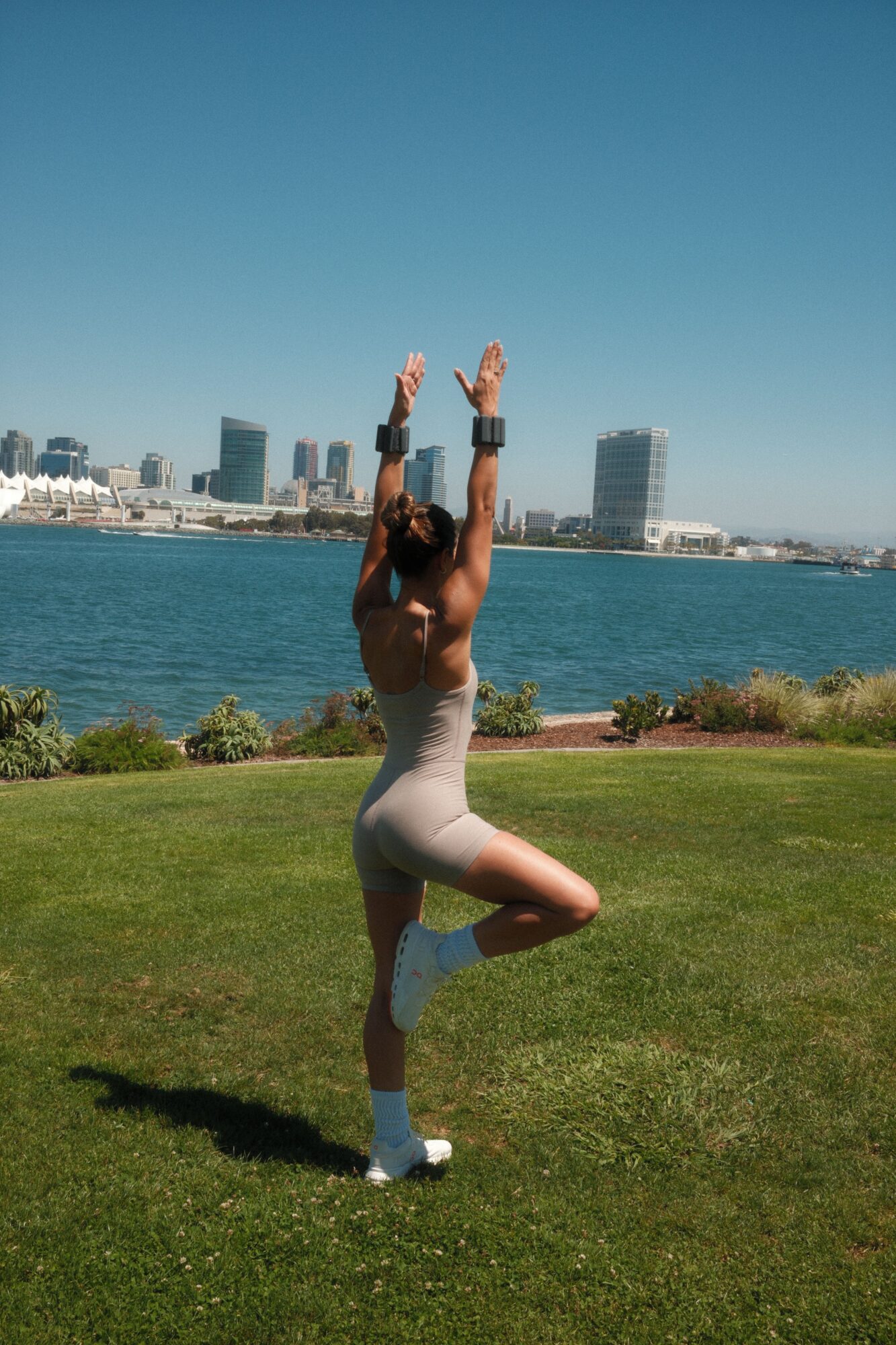Person kneeling on grass near water with city skyline in background, arms raised, sunny day.