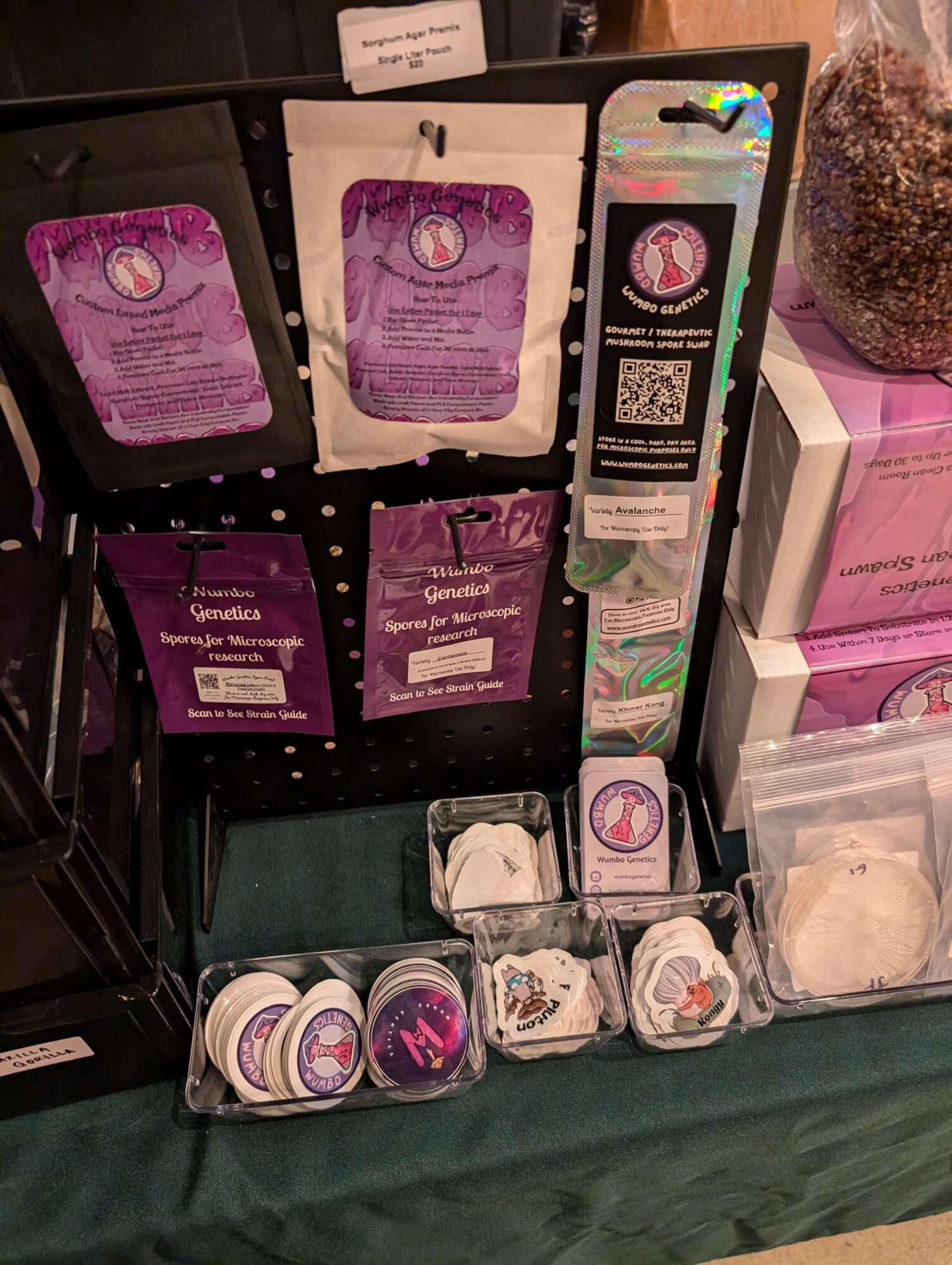 Display of purple and pink packaged products, small containers, and a QR code on a black stand, with a glass jar nearby.