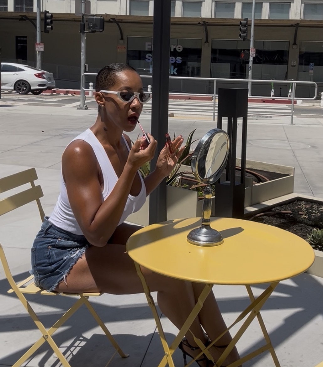 Woman sitting at yellow table applying makeup, wearing sunglasses and a white tank top, with a city street background.