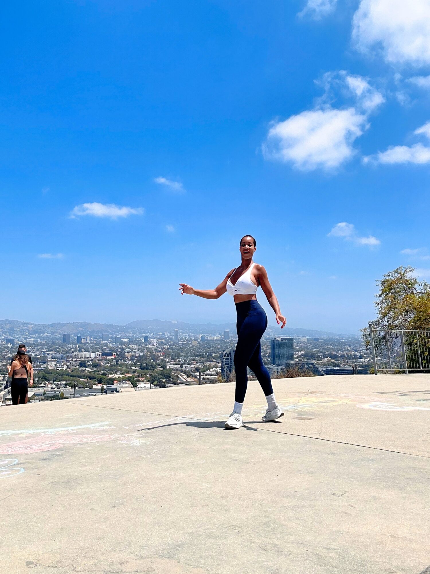 Person standing outdoors on a sunny day with a cityscape in the background, blue sky with clouds, wearing a white top and dark pants.