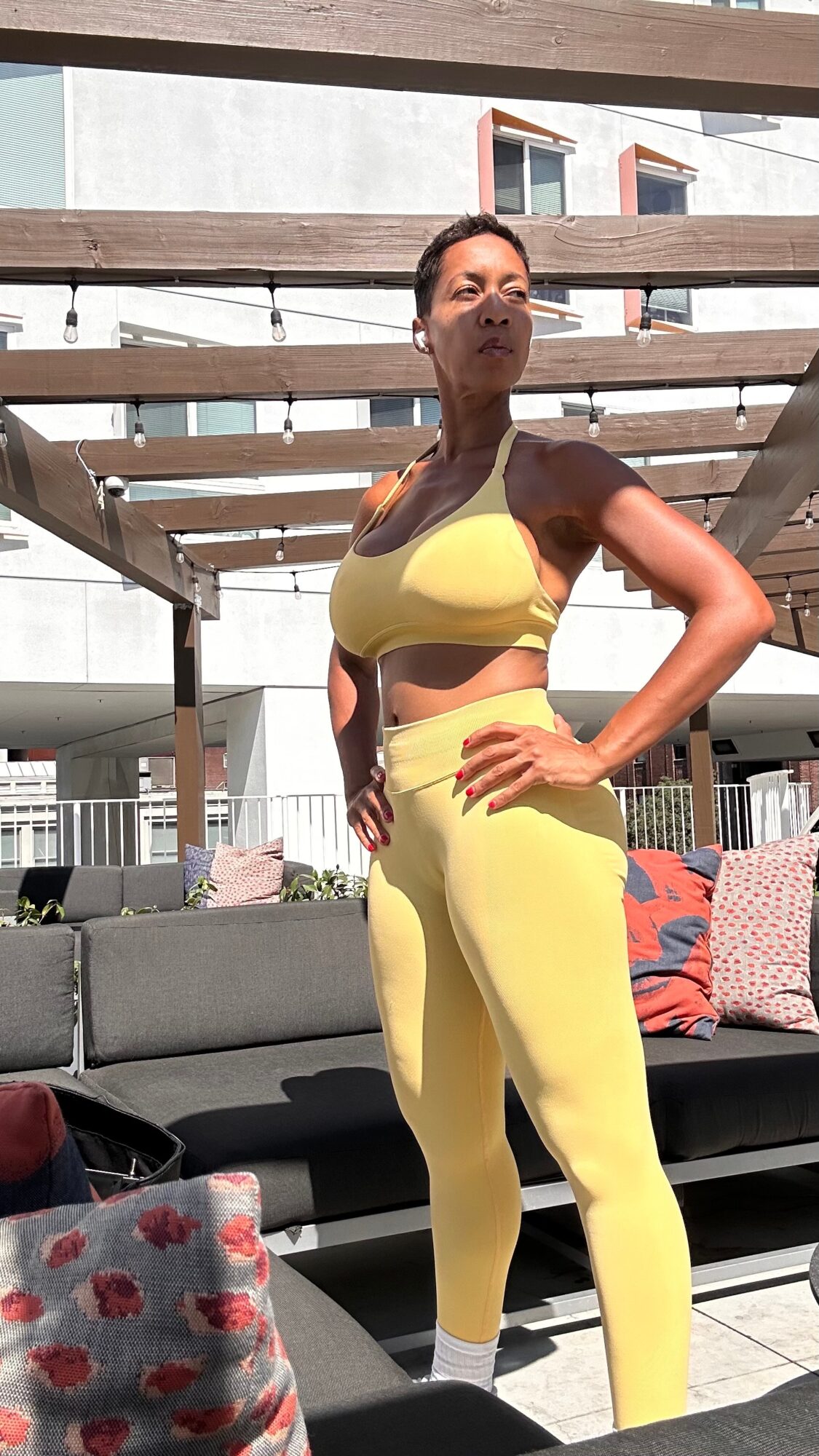 Woman in yellow workout outfit standing outdoors under a pergola with buildings in background.