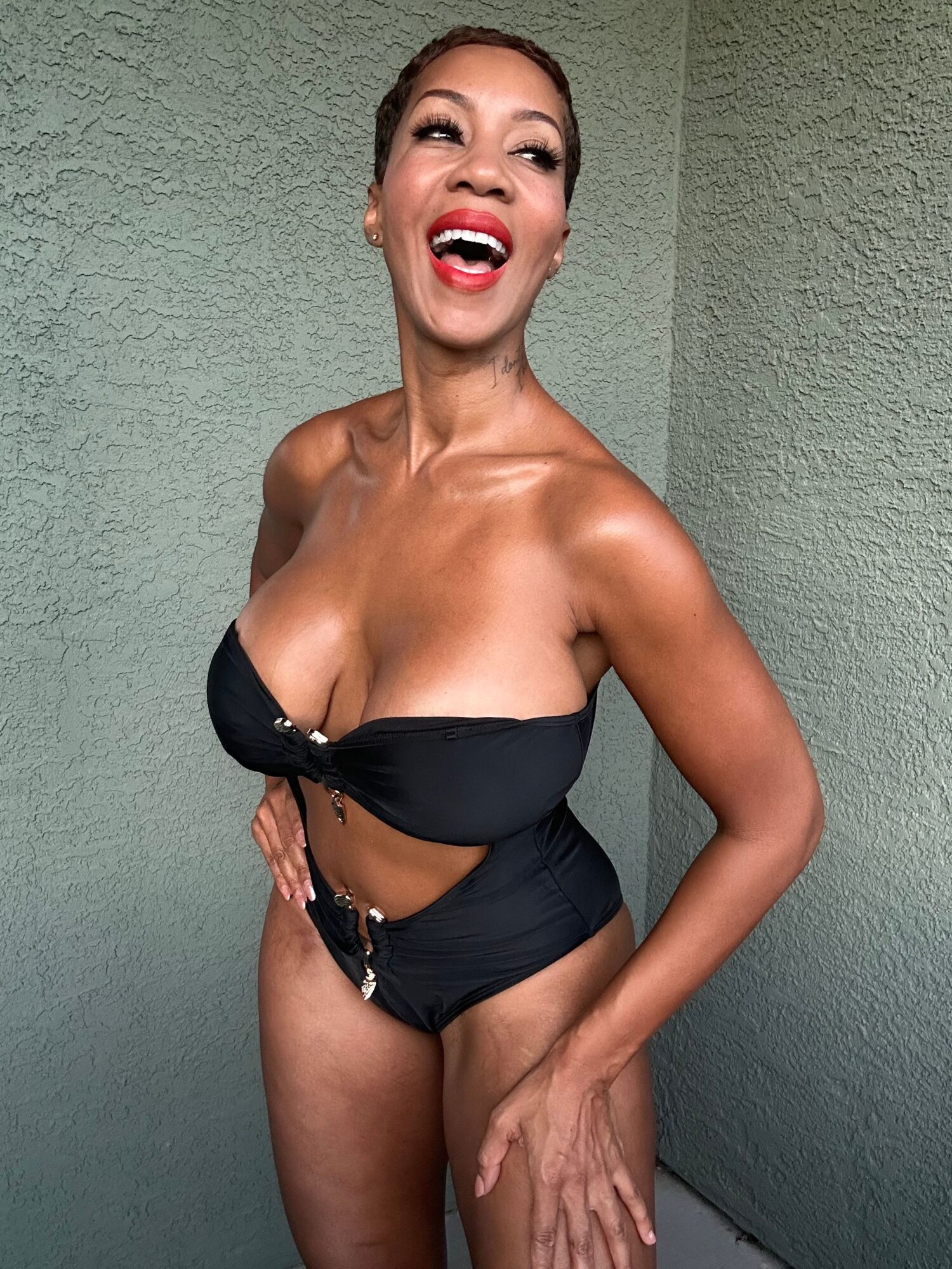 Woman with short hair, wearing a black strapless top, smiling, standing against a green wall.