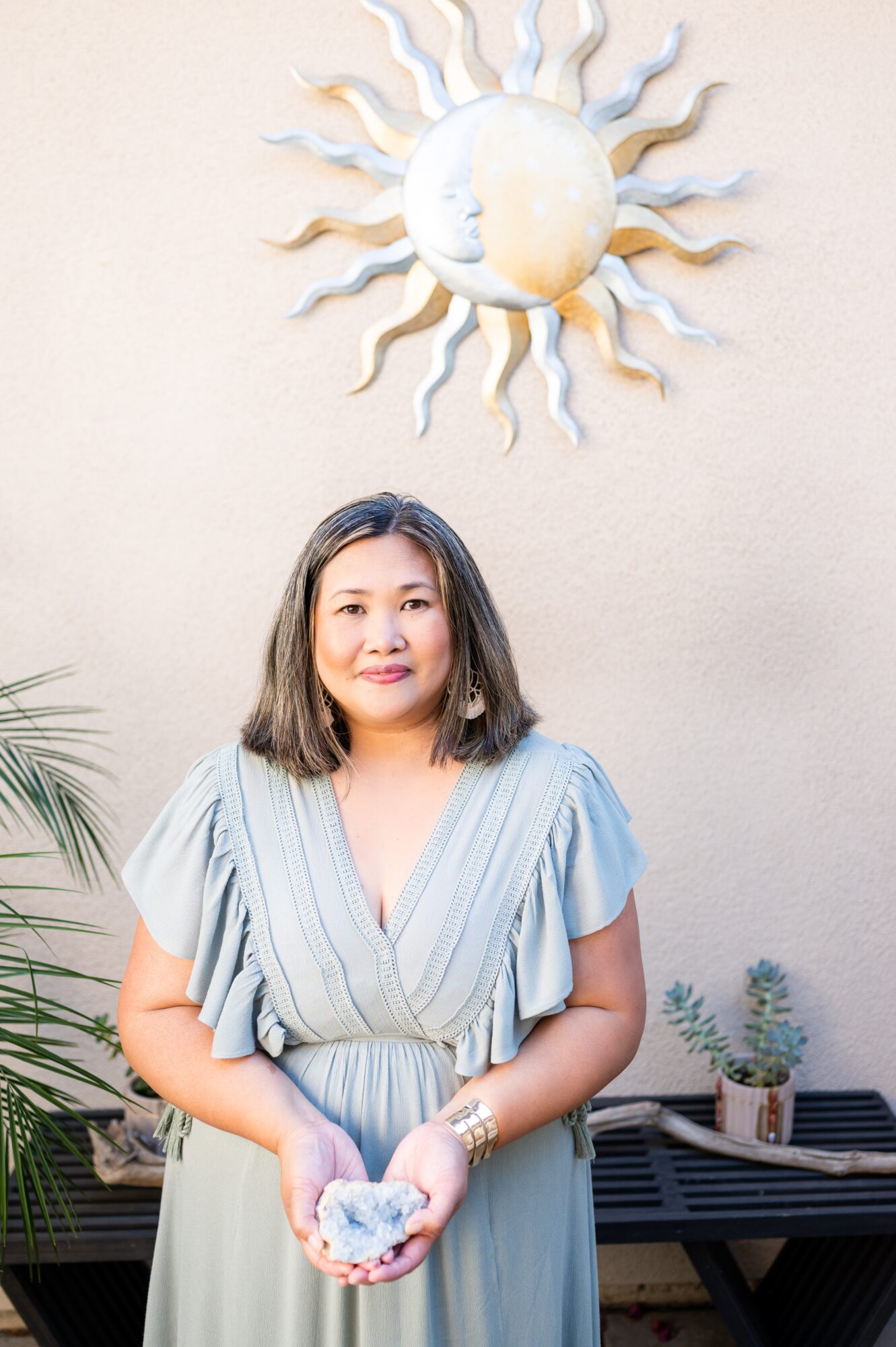 Woman with shoulder-length hair in a light blue dress holding a bowl, standing outdoors with a sun and moon wall decoration behind her.