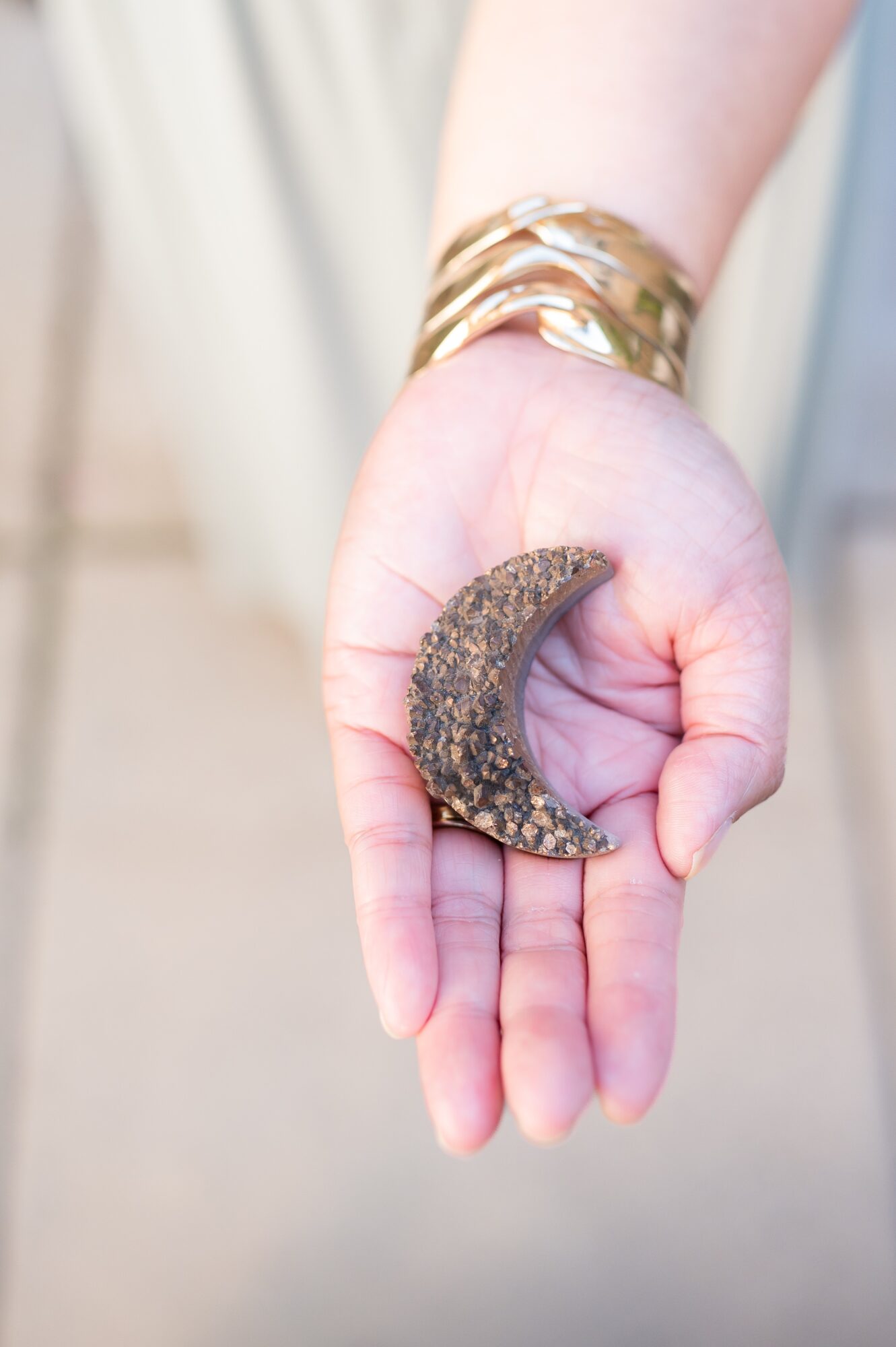 Person's hand holding a crescent-shaped object, wearing multiple gold bracelets, with a blurred background.