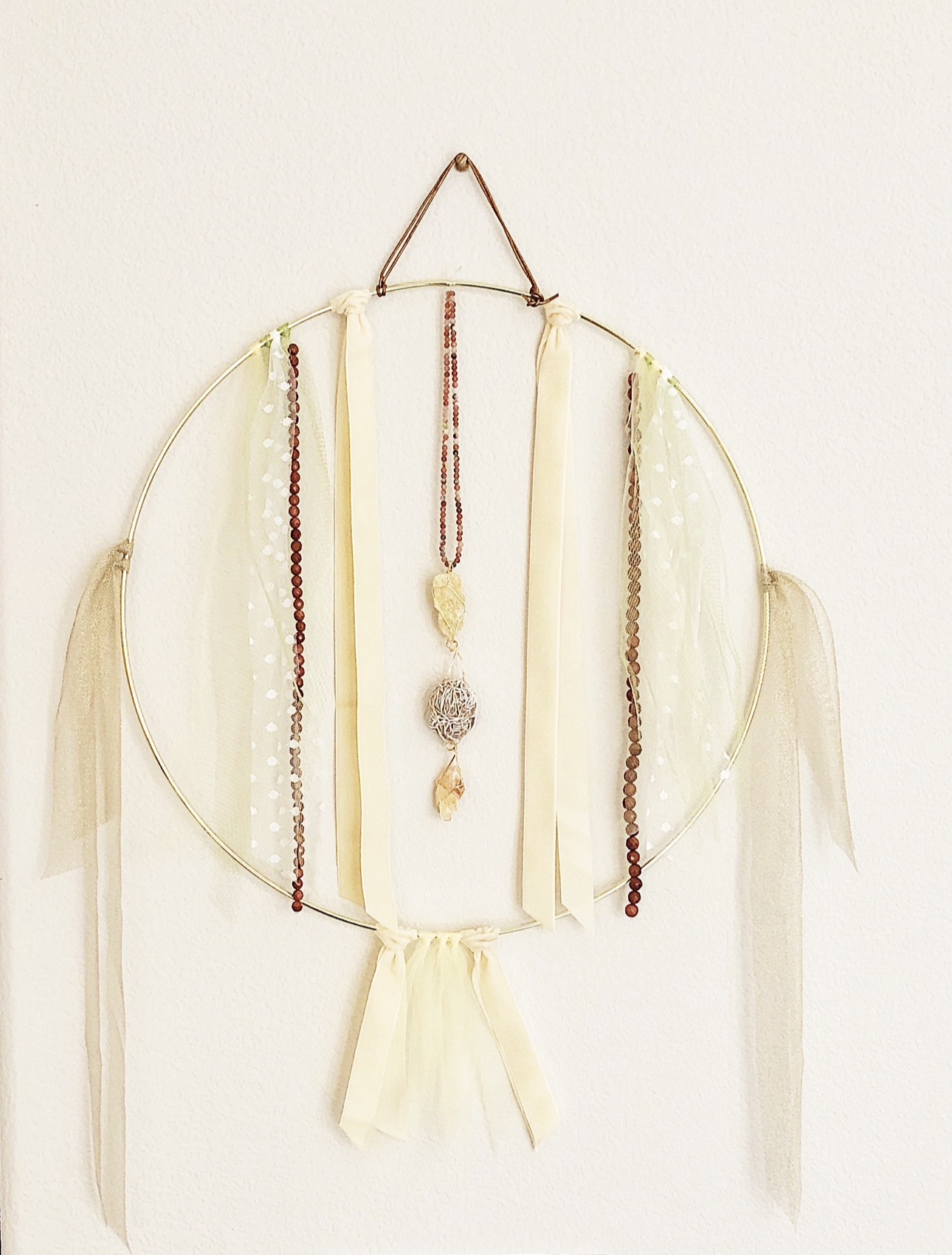 Decorative wall hanging with ribbons and beads, featuring a central fabric strip with jewelry, on a plain white background.