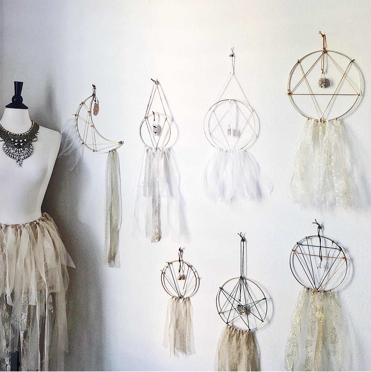 Display of eight dreamcatchers hanging on a white wall, with a mannequin dressed in a beige skirt and necklace on the left.