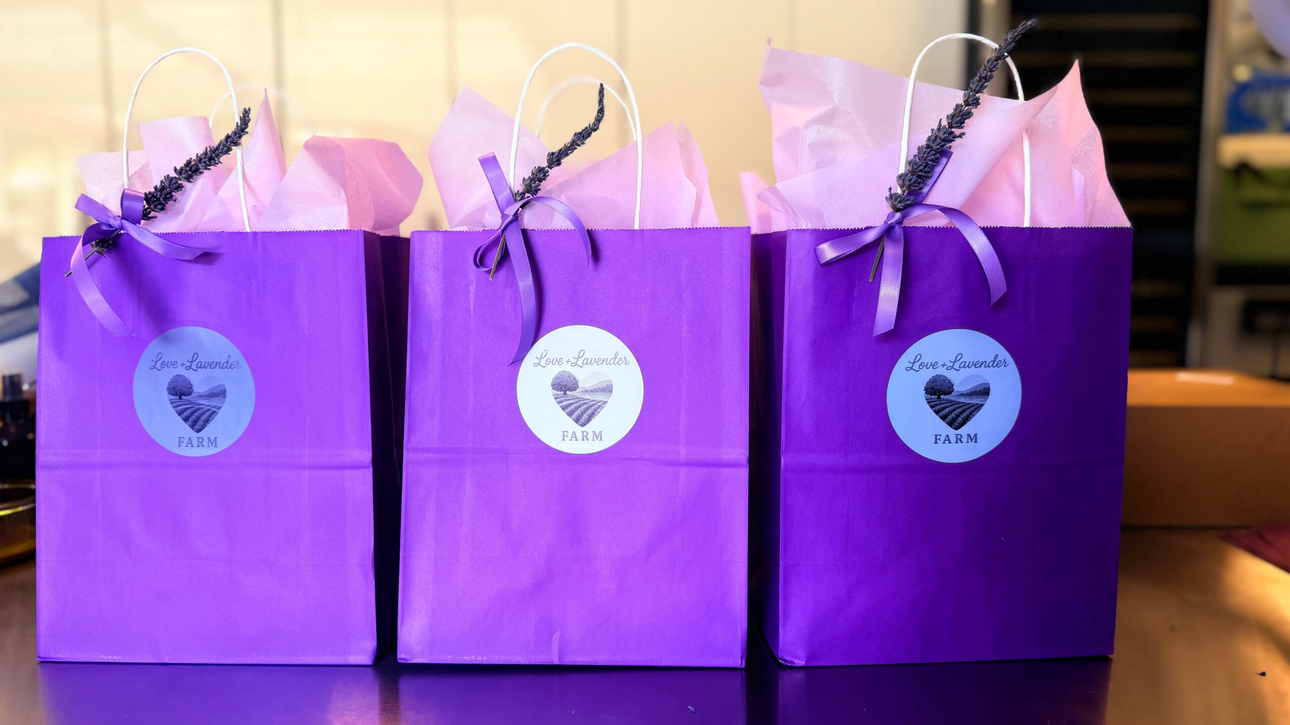 Three purple gift bags with heart-shaped stickers and ribbons, placed on a surface.
