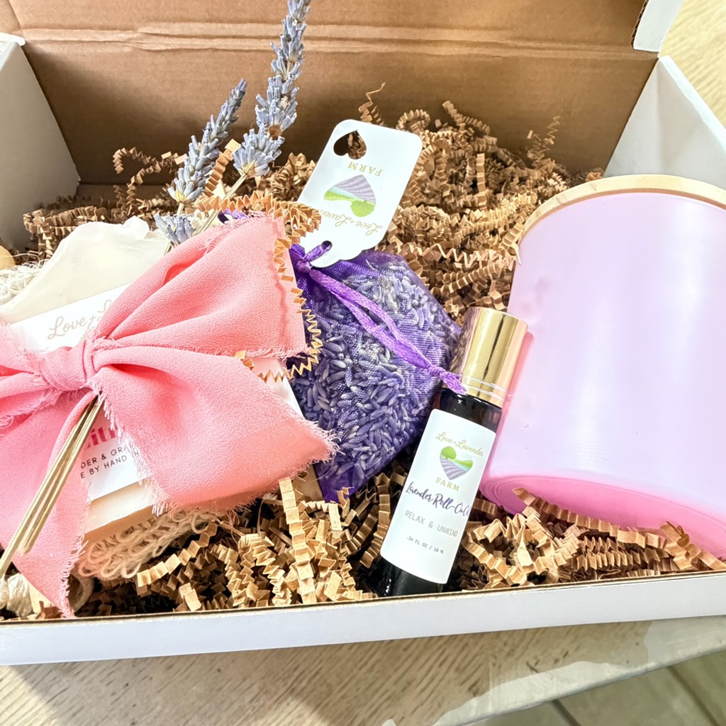 Open box with pink bow, purple lavender, small bottle, and pink container, surrounded by shredded paper and decorative lavender sprigs.