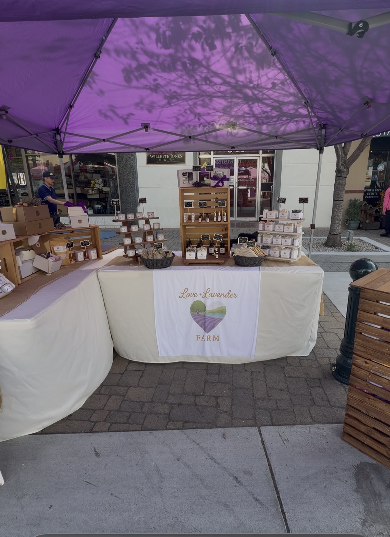 Market stall with various products under a purple canopy, tablecloth with a heart logo, on a city sidewalk.