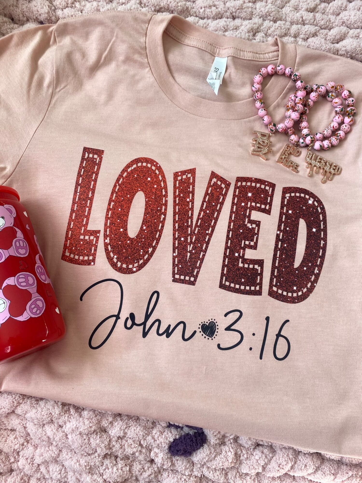 Beige shirt with red and black text, pink beaded bracelets, and a red object partially visible on a textured surface.