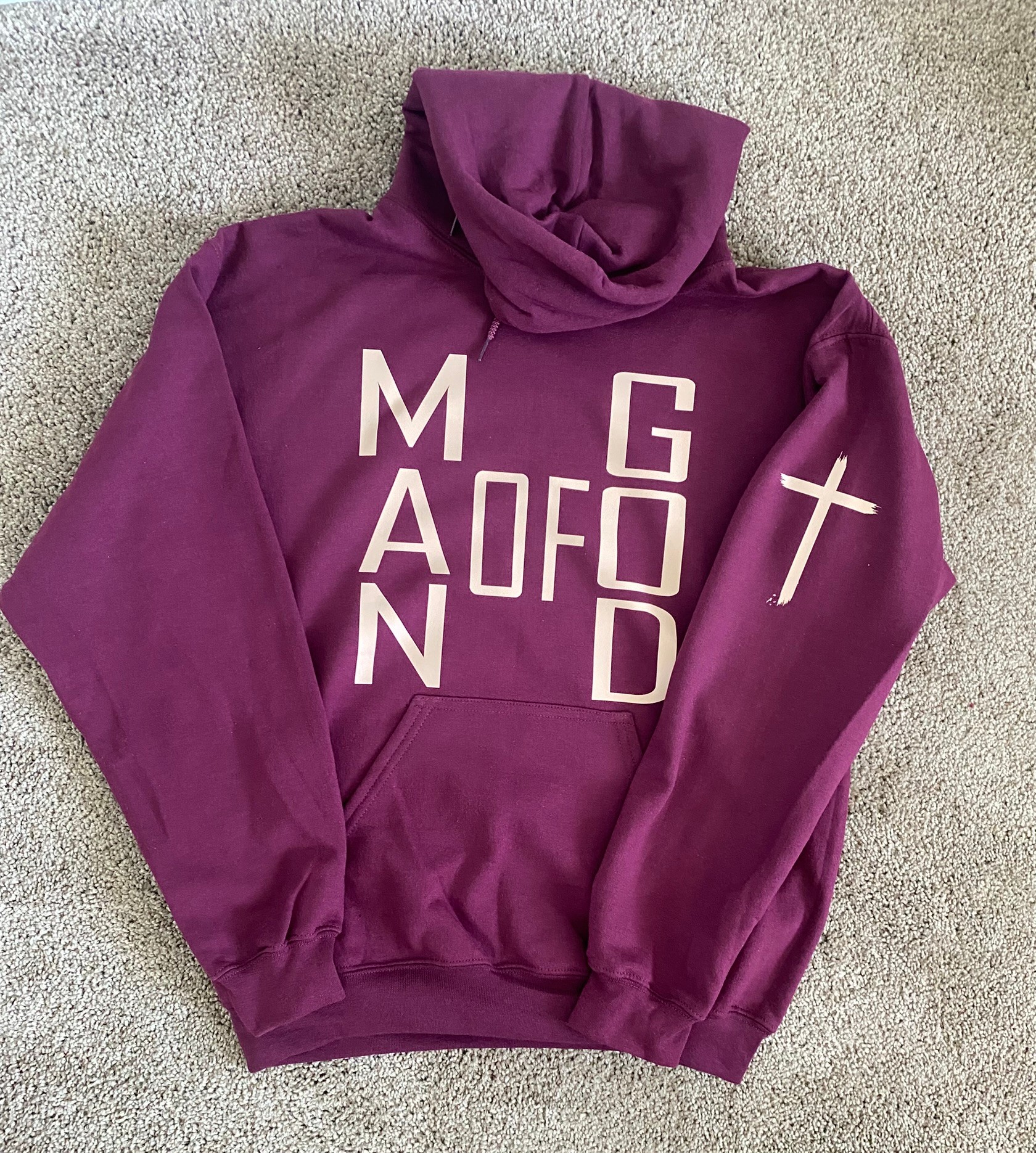 Purple hoodie with white letters and a cross symbol on the sleeve, laid flat on a gray surface.