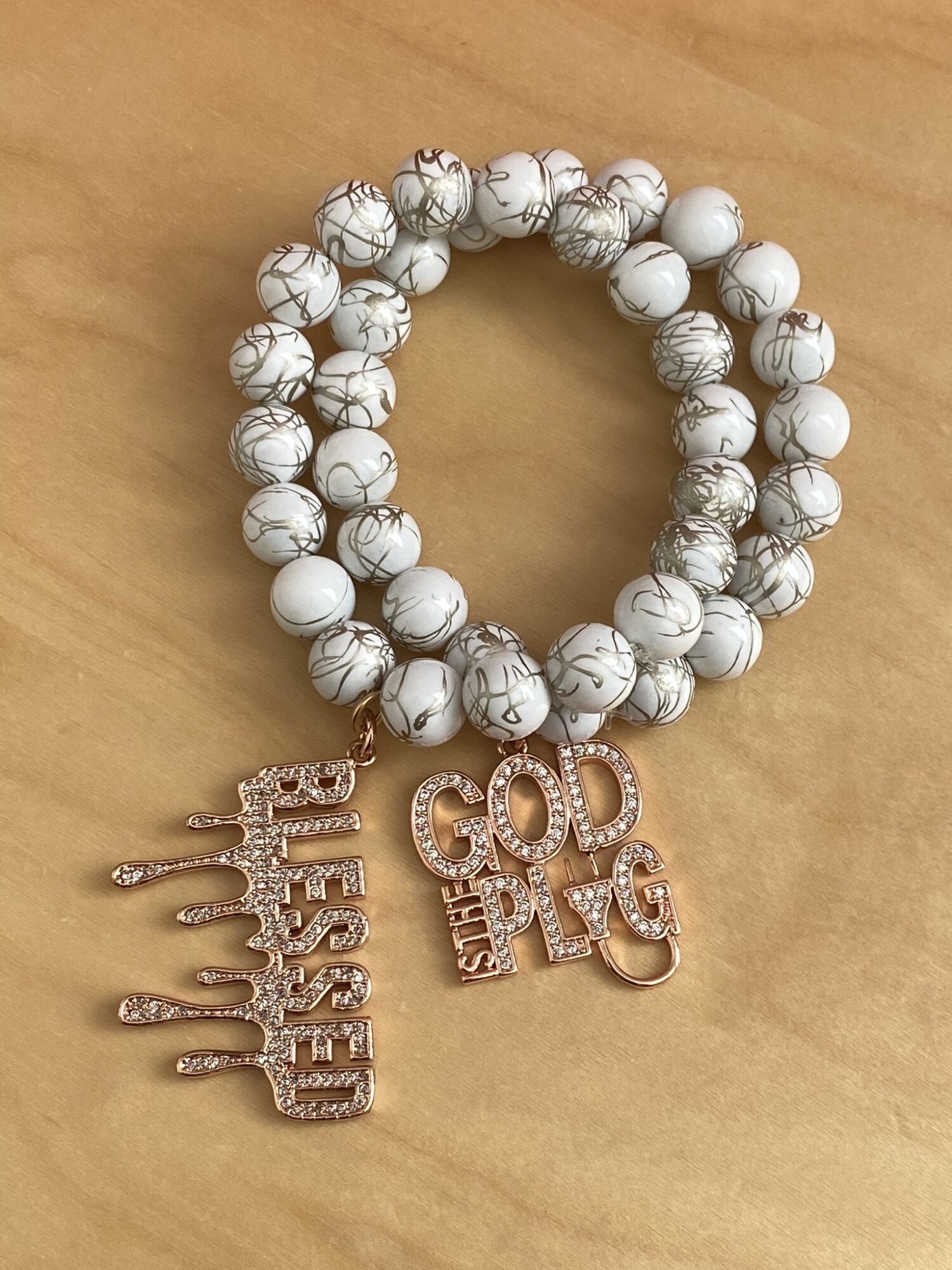 Two beaded bracelets with white and gray marbled beads and gold-colored charms reading 'Blessed' and 'God is Love' on a wooden surface.