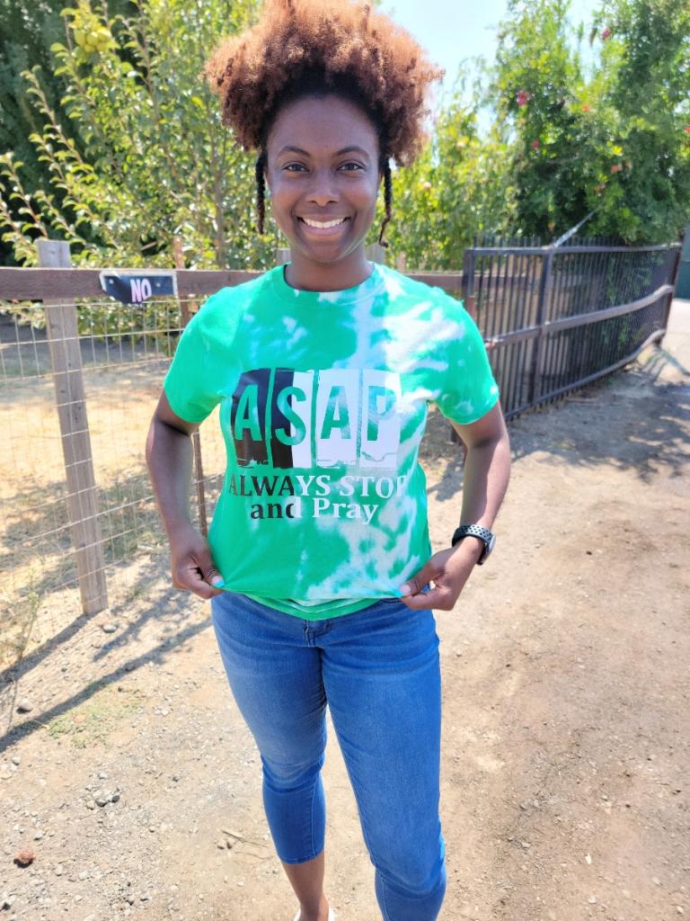 Young woman with curly hair smiling outdoors, wearing a green and white tie-dye shirt and blue jeans.