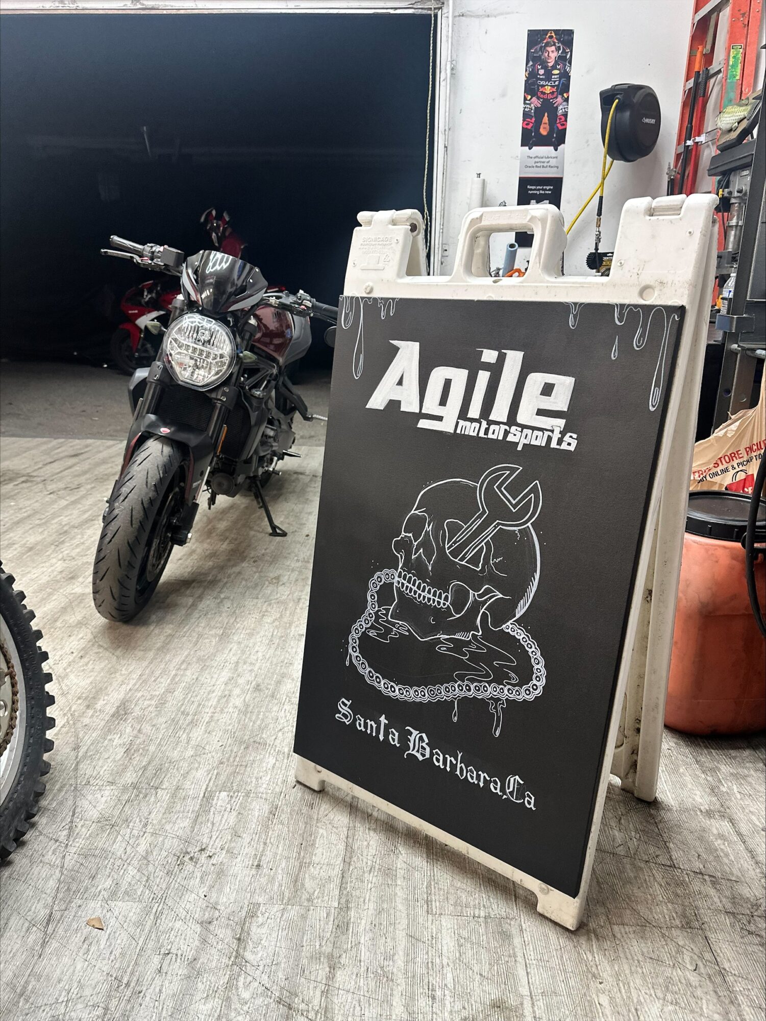 A blackboard sign with white text and a drawing, placed on a wooden floor near a motorcycle inside a garage or shop.