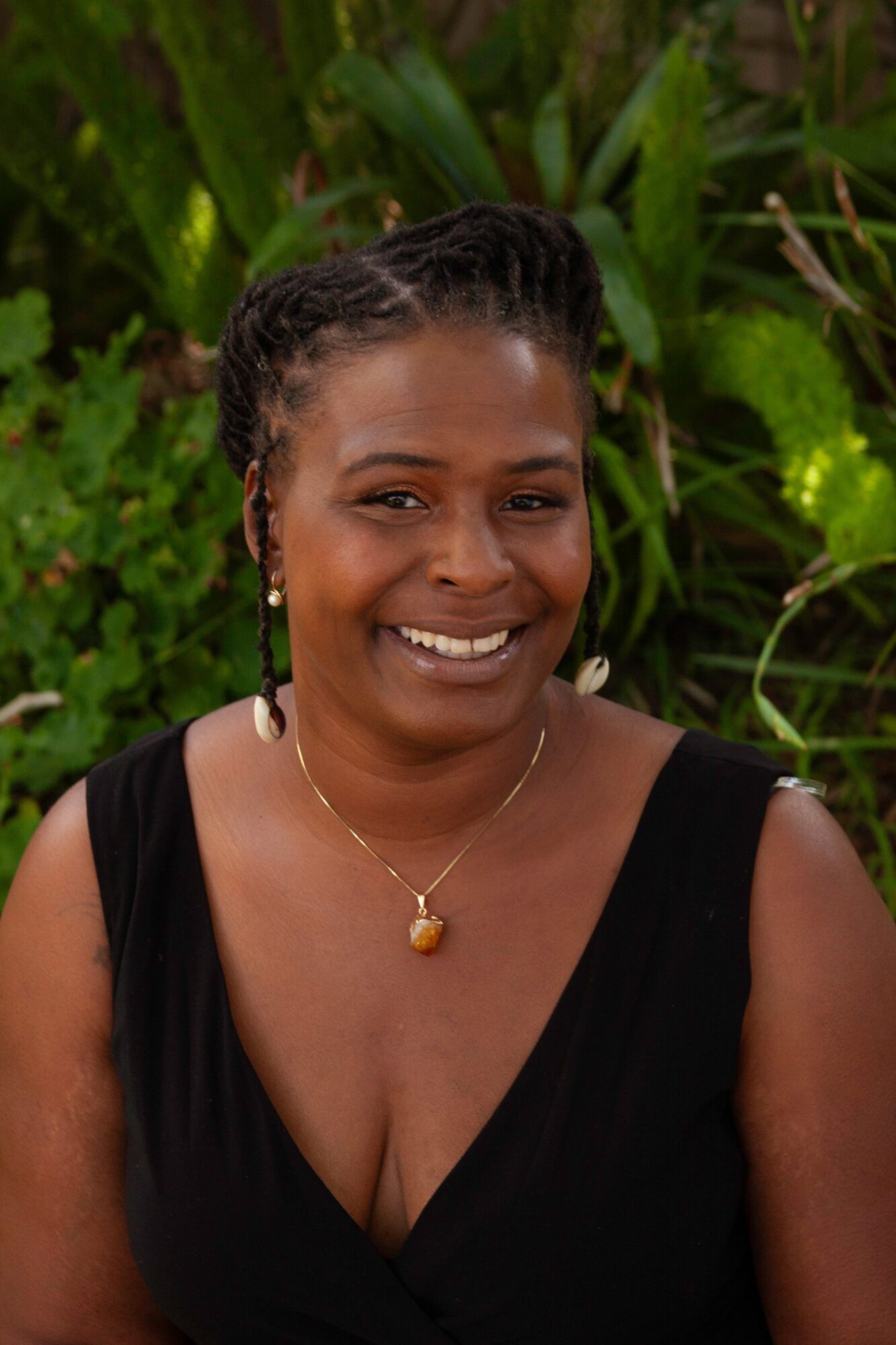 Smiling woman with braided hair, wearing a black dress and gold jewelry, outdoors with green plants in background.