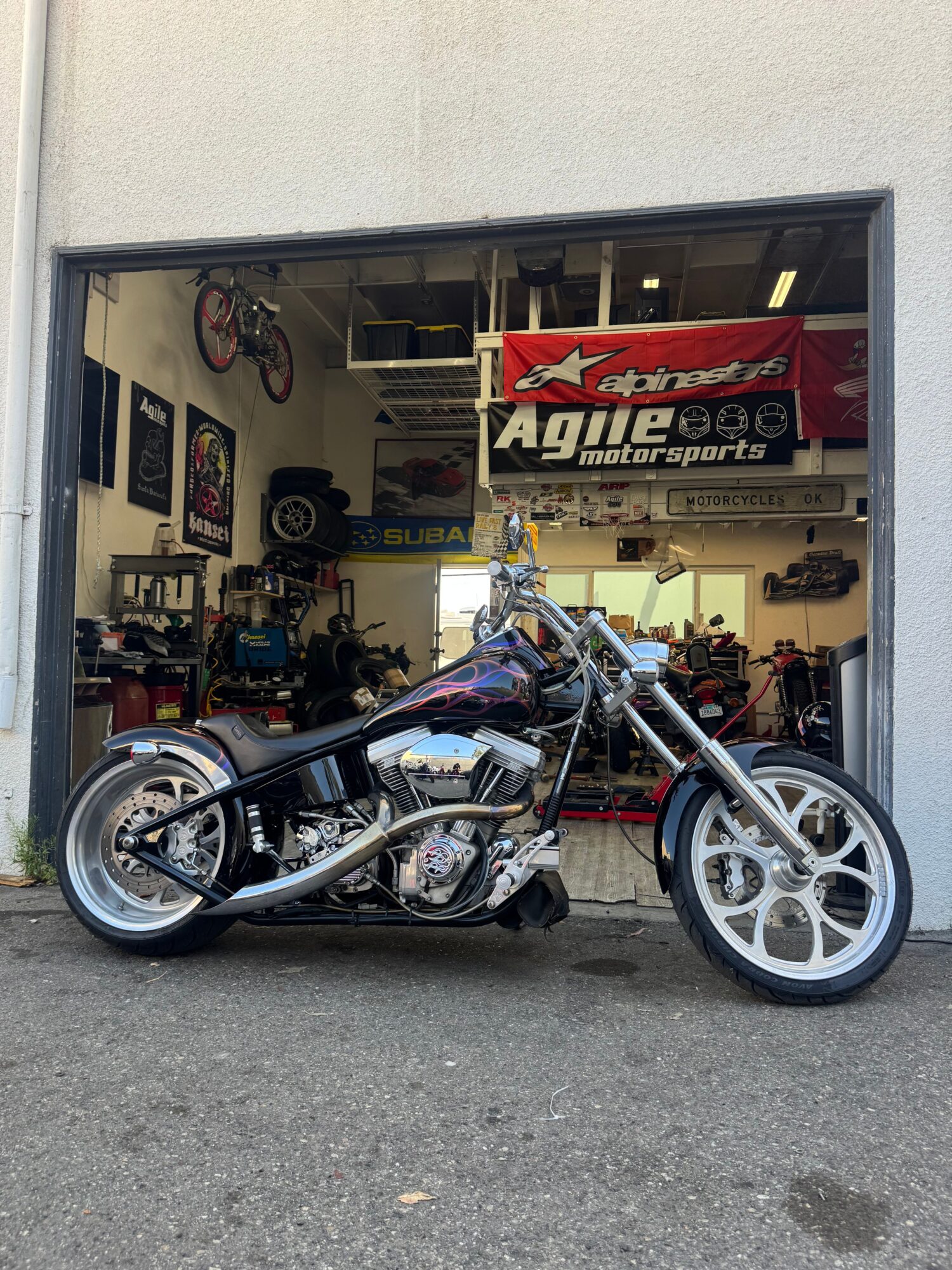Motorcycle inside a garage with various tools and equipment, viewed from the side, with a wheel removed and leaning on a stand.