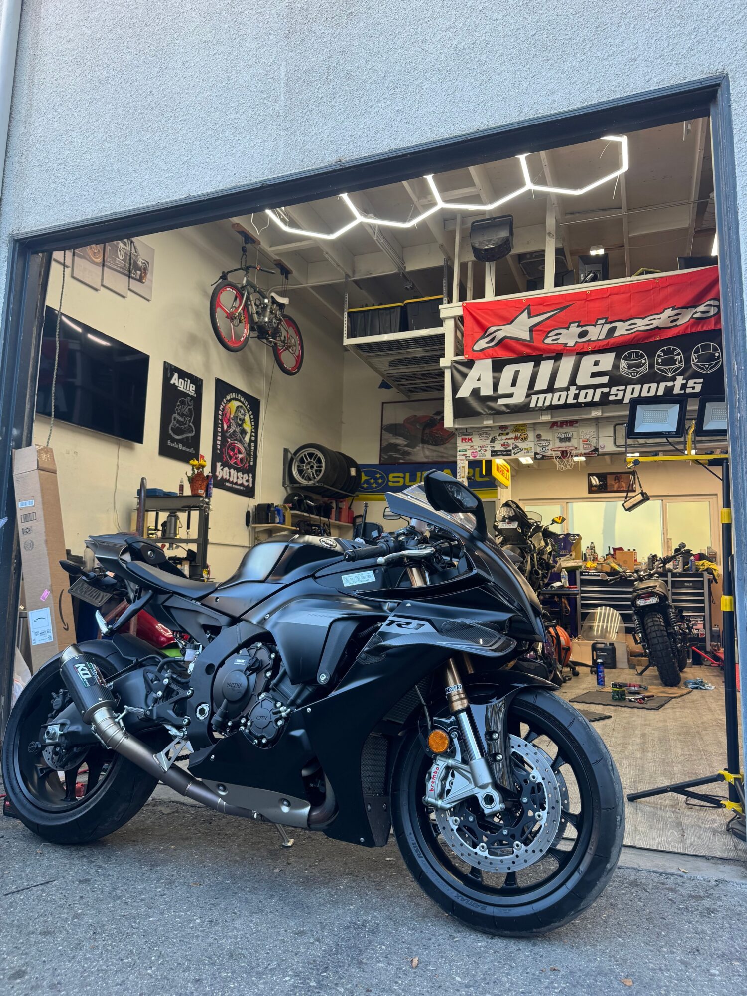 Black motorcycle parked inside a garage with tools and equipment, viewed from the side, with a sign in the background.