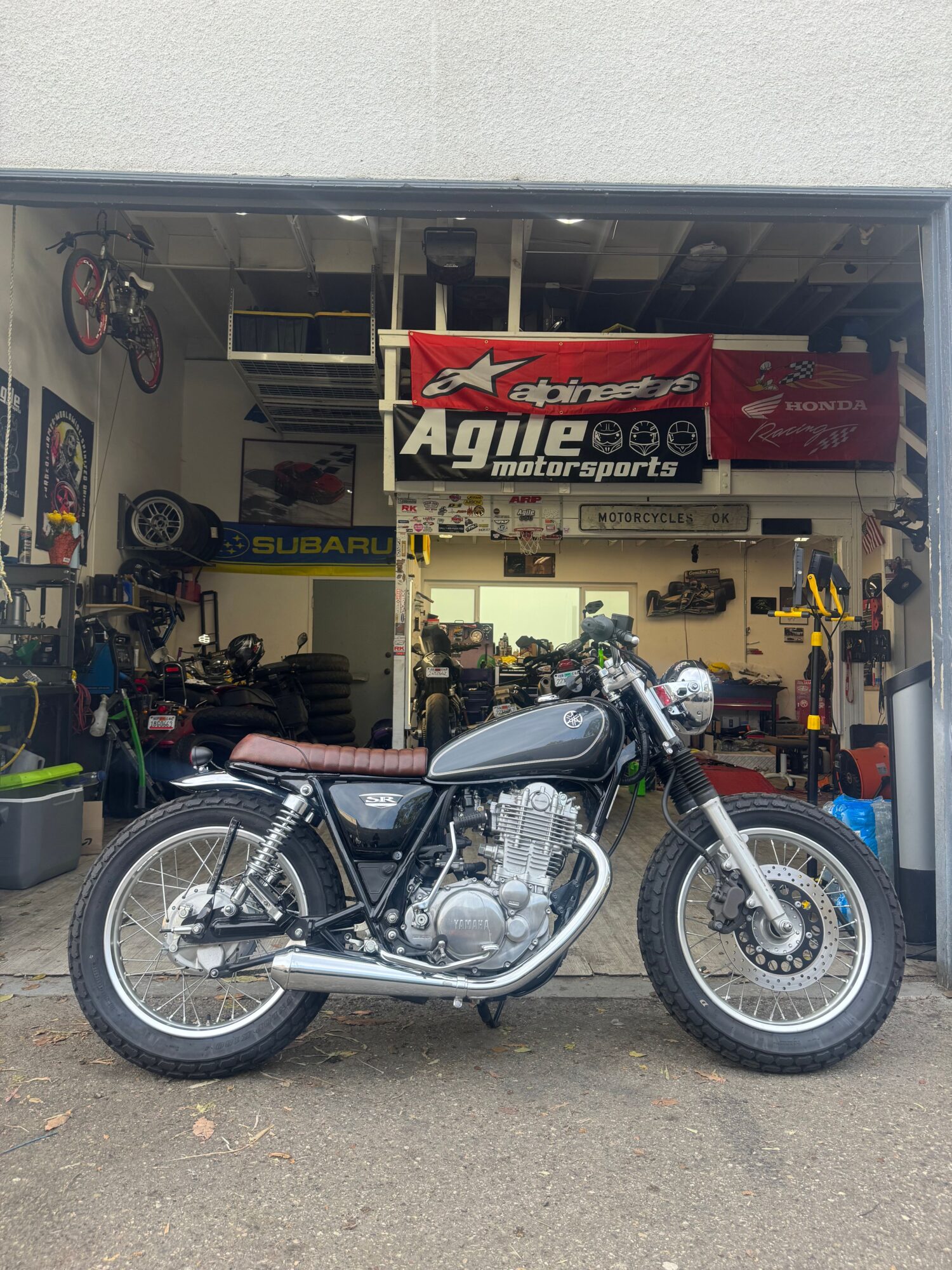 Motorcycle parked outside a garage with tools and bikes inside, viewed from the side.