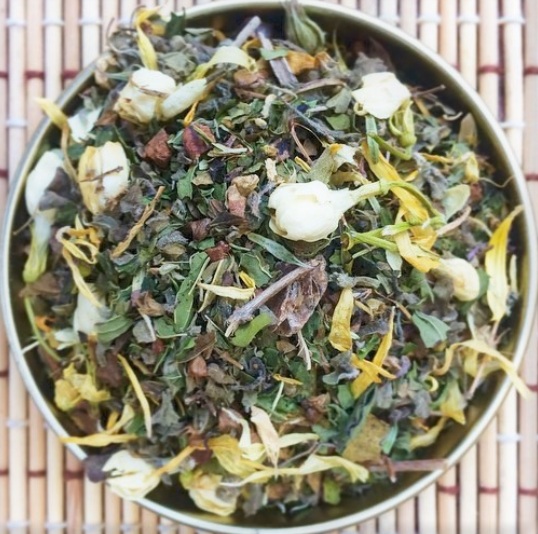 A bowl of mixed dried herbs and flowers on a bamboo mat, with green, yellow, and purple elements.