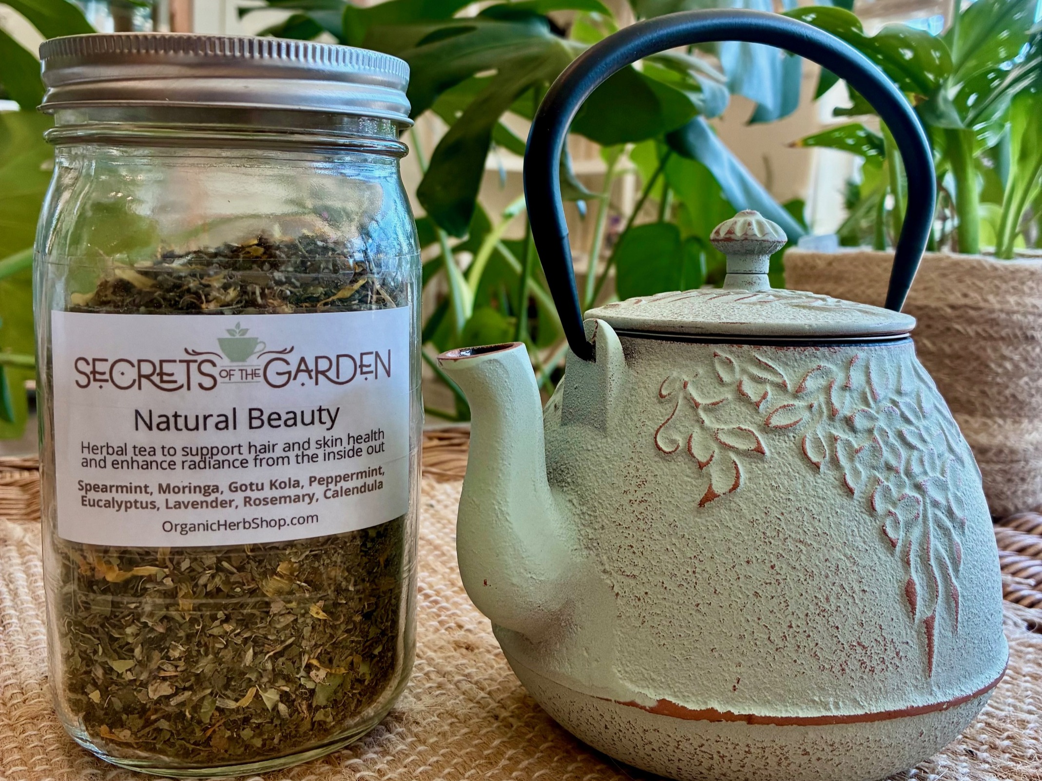 Jar of herbal tea next to a white teapot with a textured floral design, on a woven surface with green plants in background.