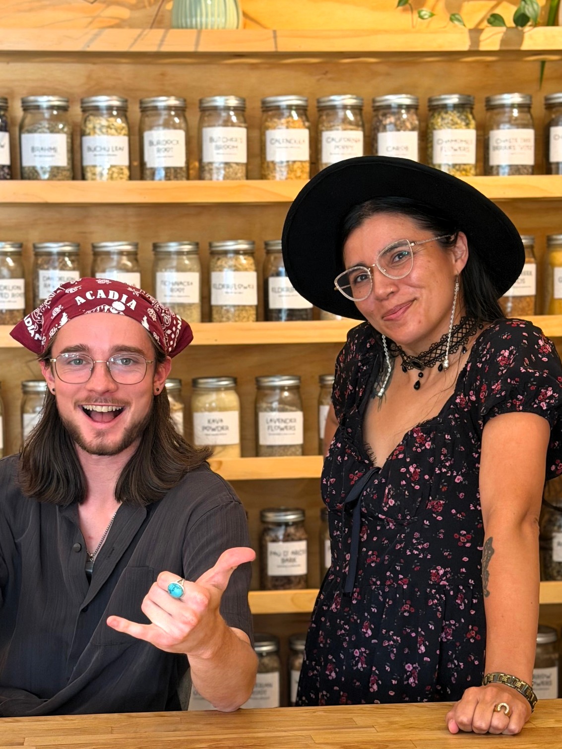 Two women standing in front of shelves filled with jars, smiling, one wearing glasses and a hat, the other with a wide-brimmed hat.