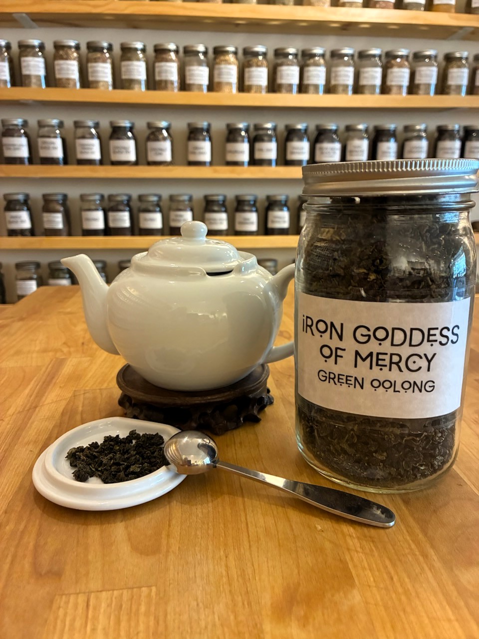 Jar labeled 'Iron Goddess of Mercy Green Oolong' tea leaves, teapot, small dish with tea leaves, spoon, and shelves of jars in background.