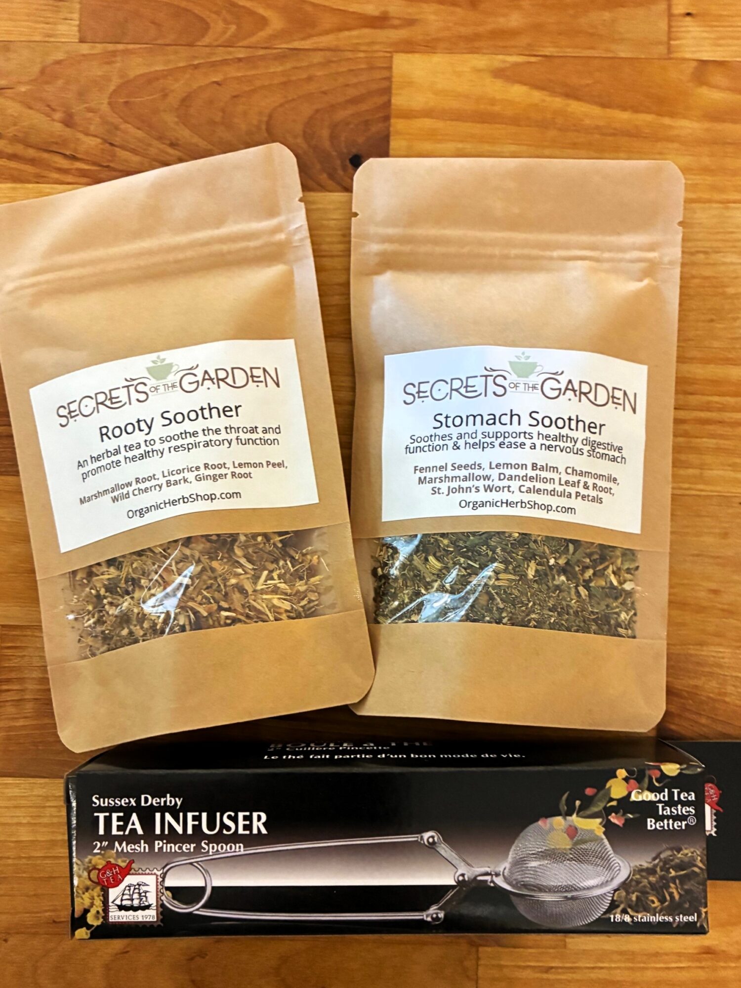 Two beige resealable bags of herbal teas labeled 'Rooty Soother' and 'Stomach Soother' on a wooden surface, with a tea infuser box below.