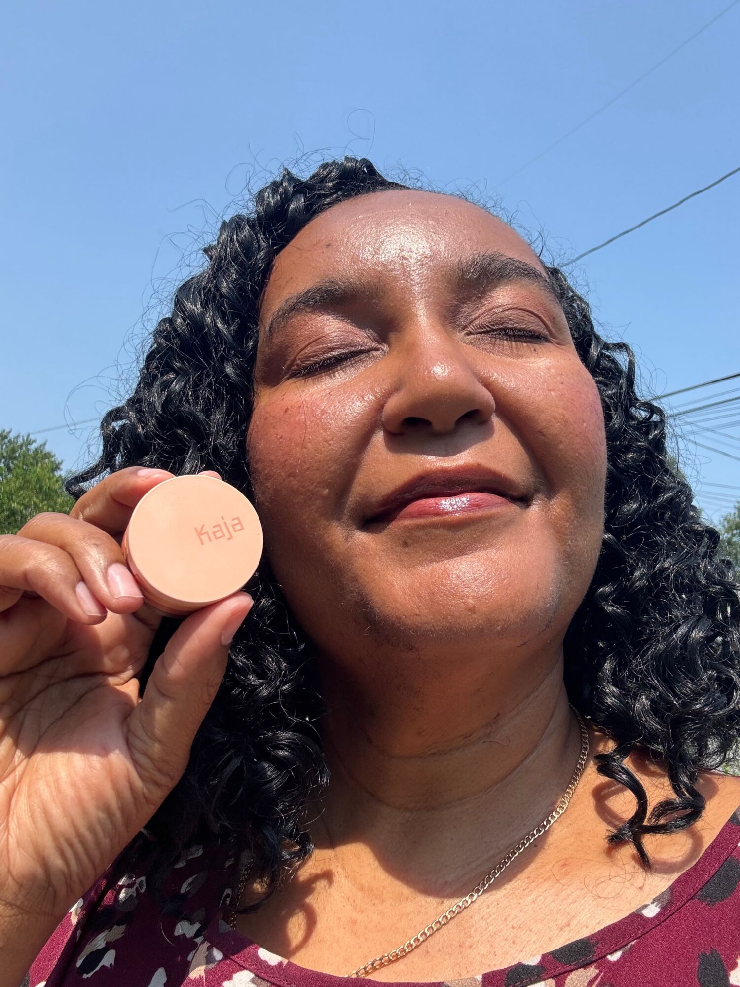 Woman with curly hair smiling outdoors, holding a small round object near her face, blue sky background.