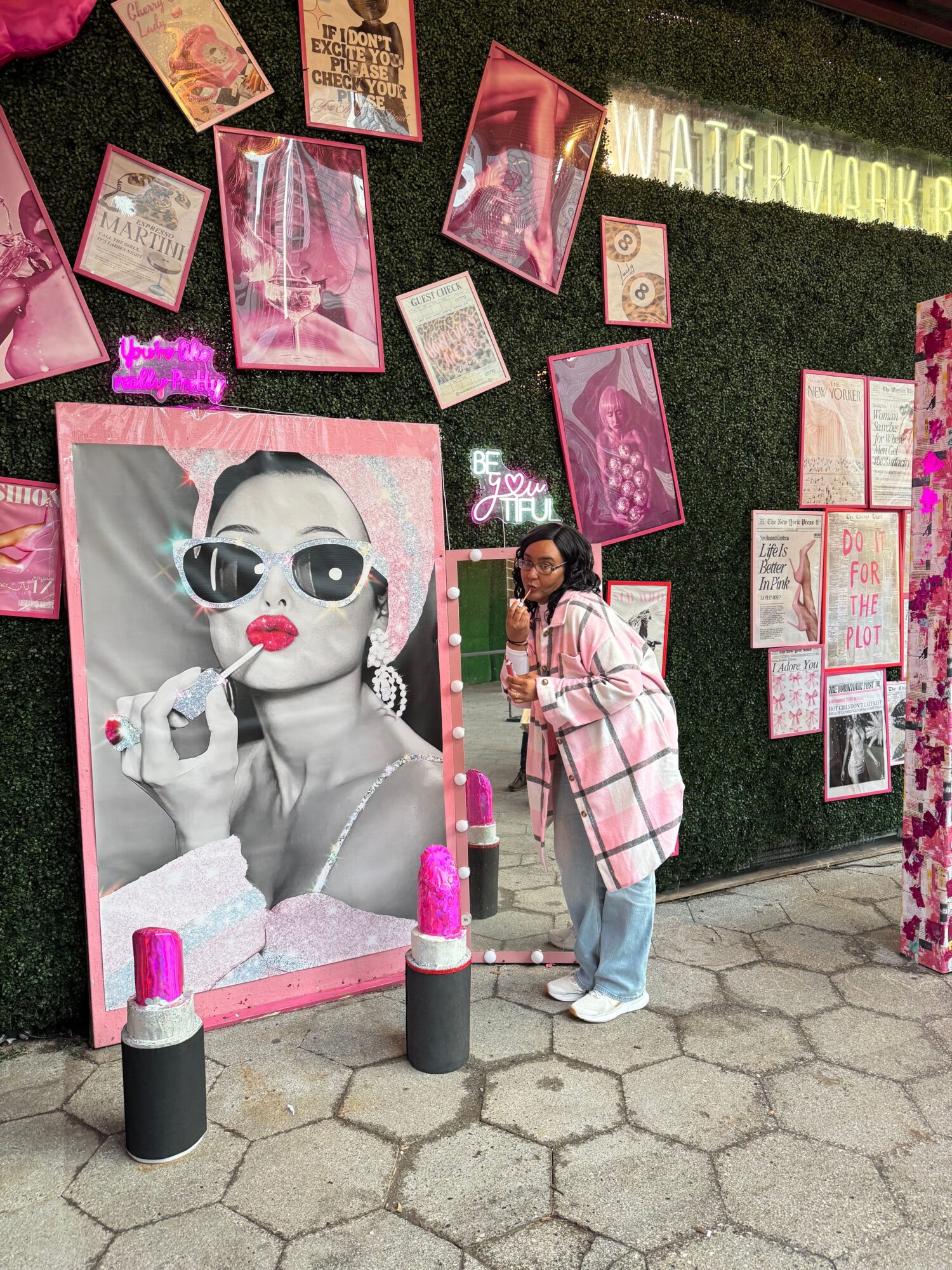 Person with pink hair and glasses standing near a large black-and-white portrait of a woman with sunglasses, surrounded by pink and red decorations.