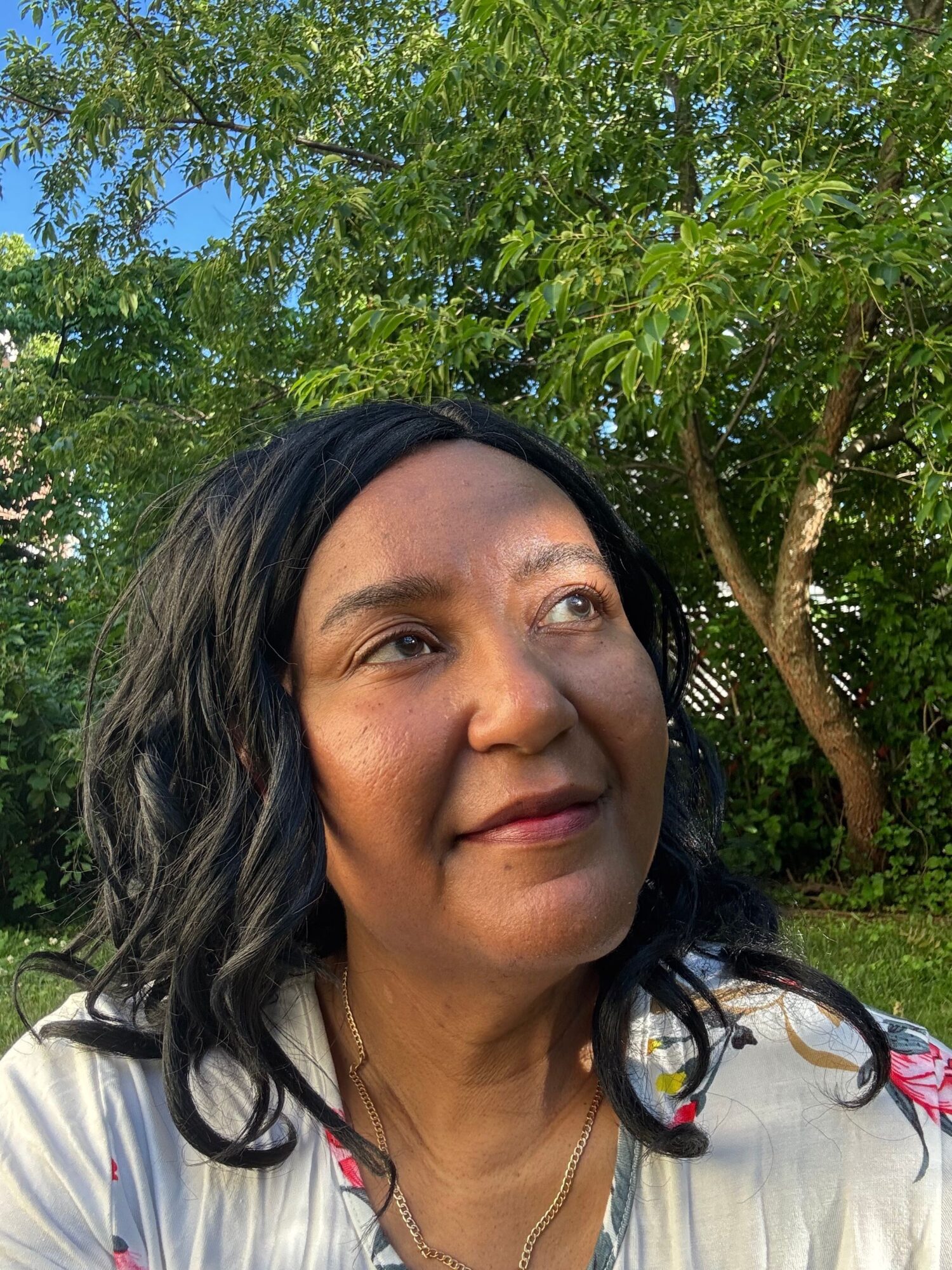 Woman with black hair and a necklace outdoors, trees and blue sky in background.
