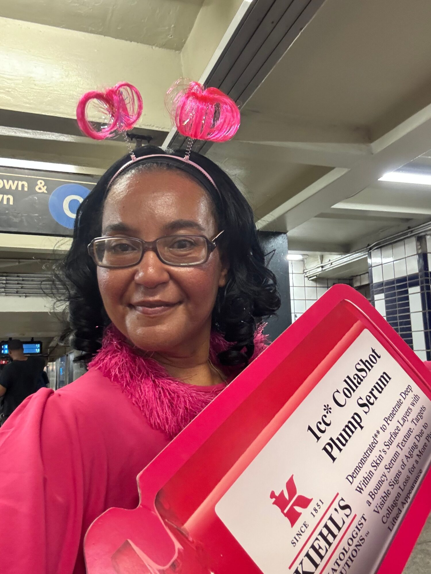 Woman with glasses and curly hair wearing a pink top, holding a pink folder, standing in a store aisle.