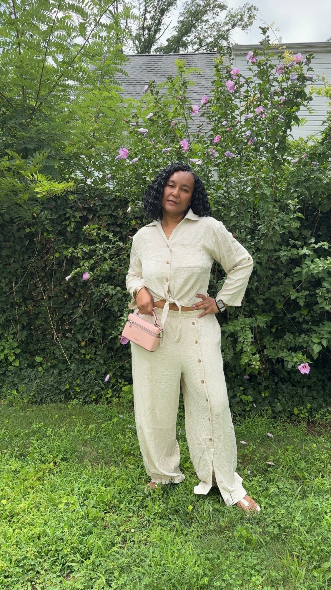 Woman standing outdoors in front of greenery and flowering bushes, wearing a cream-colored outfit and holding a pink purse.