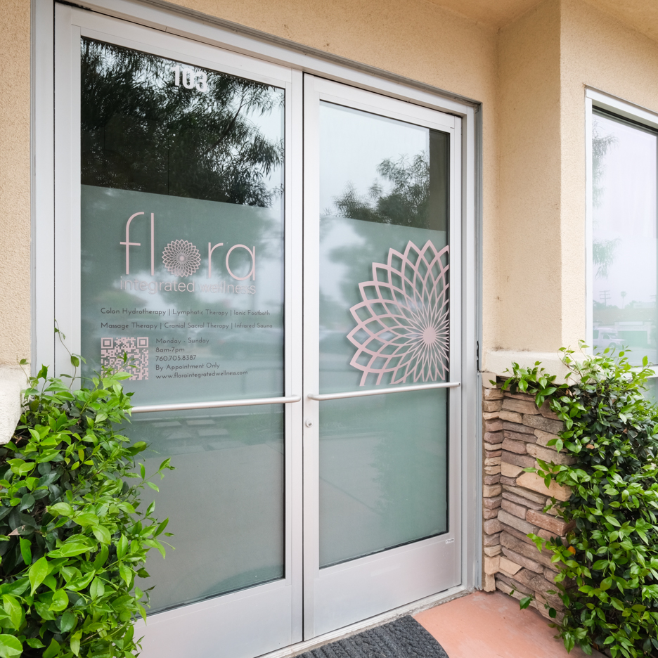 Glass door with floral design, surrounded by green bushes, with a brick wall on the right, and a reflection of trees in the glass.