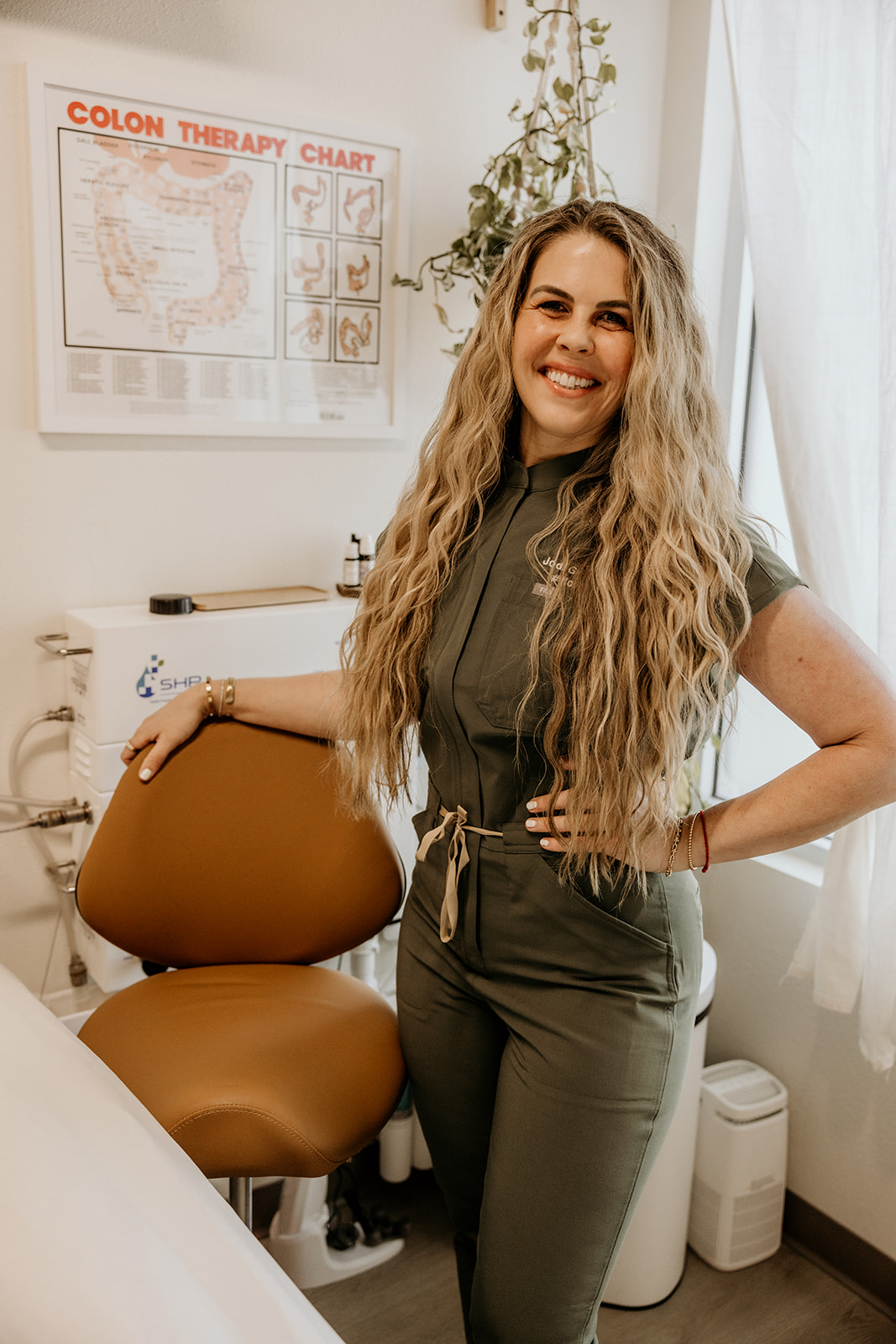 Smiling woman with long wavy hair in medical office, standing next to a brown chair, with a colon therapy chart on the wall.