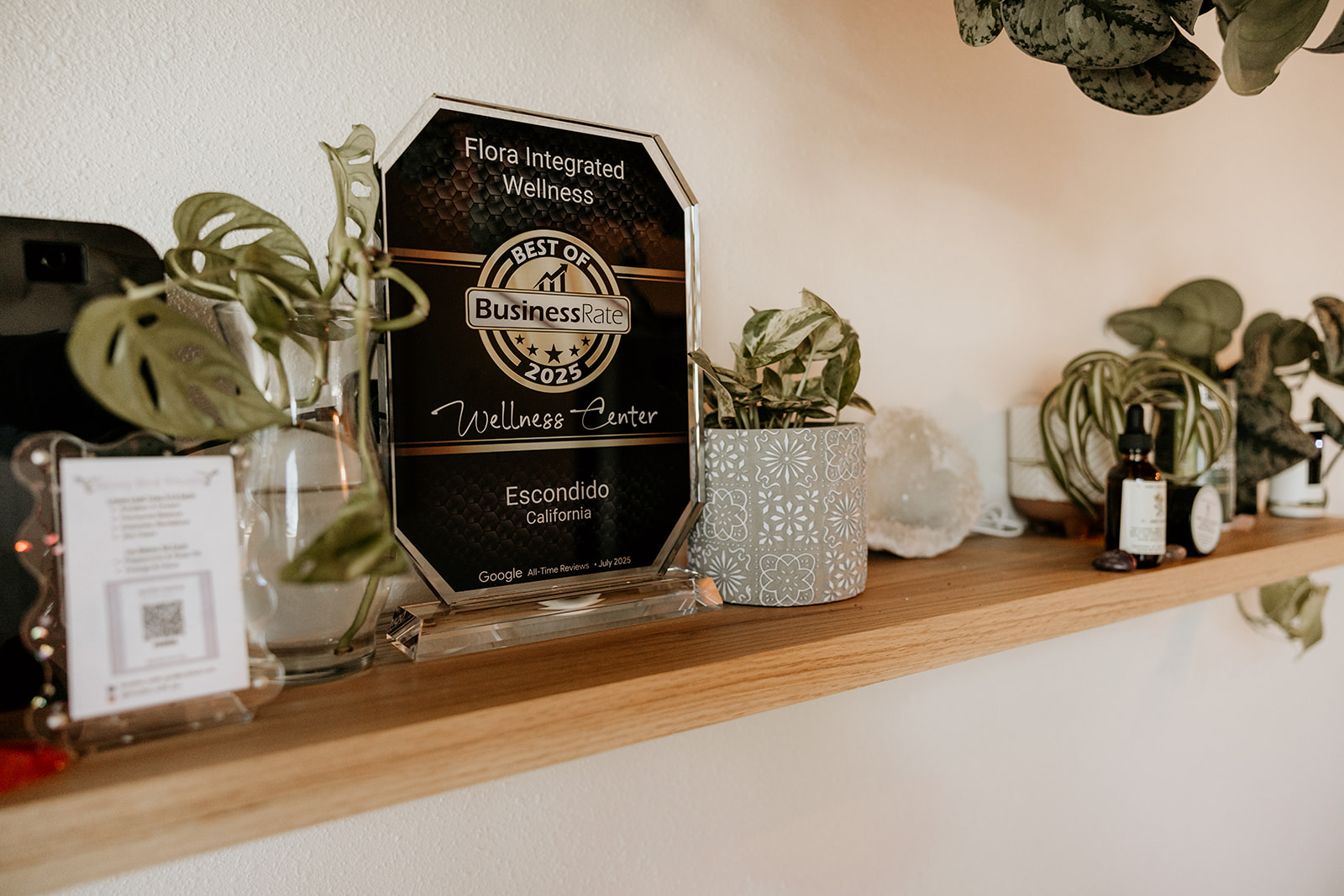 Wooden shelf with plants, a sign, and small bottles, against a plain wall.
