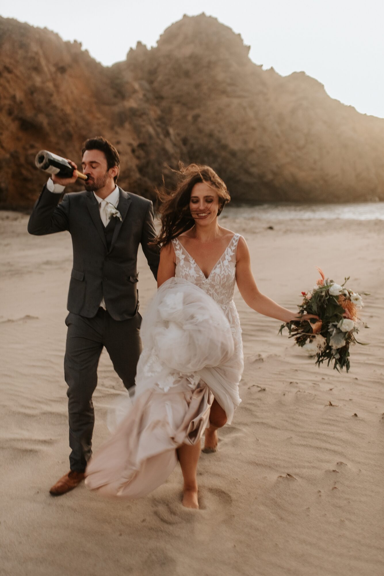 A bride and groom running on the beach, the bride holding a bouquet, the groom drinking from a bottle, with rocky cliffs in background.