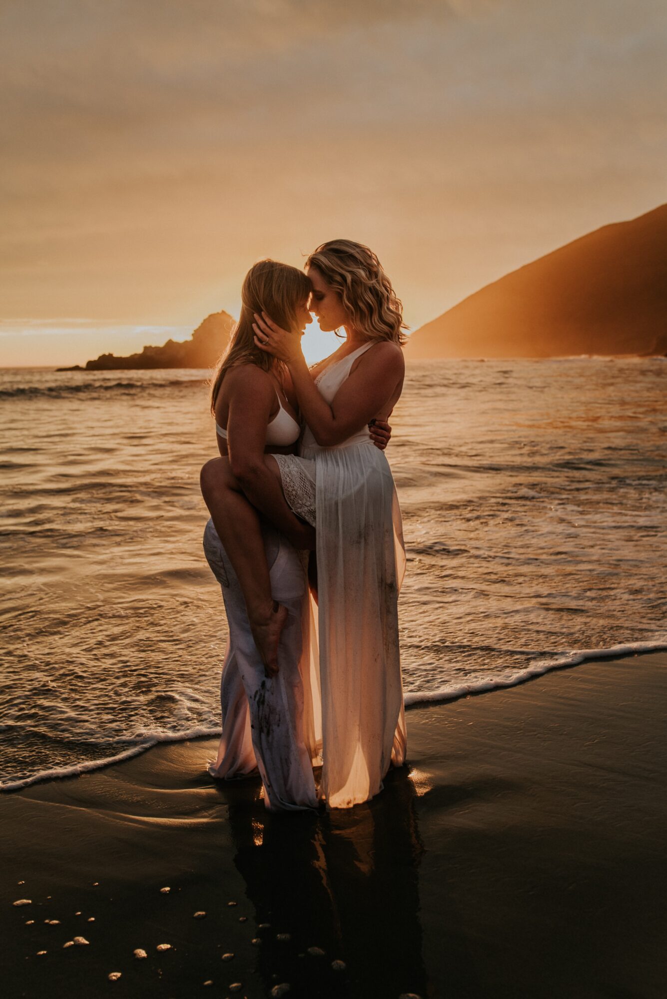 Two women standing in shallow water at sunset, embracing and touching foreheads, with a beach and sand dunes in background.