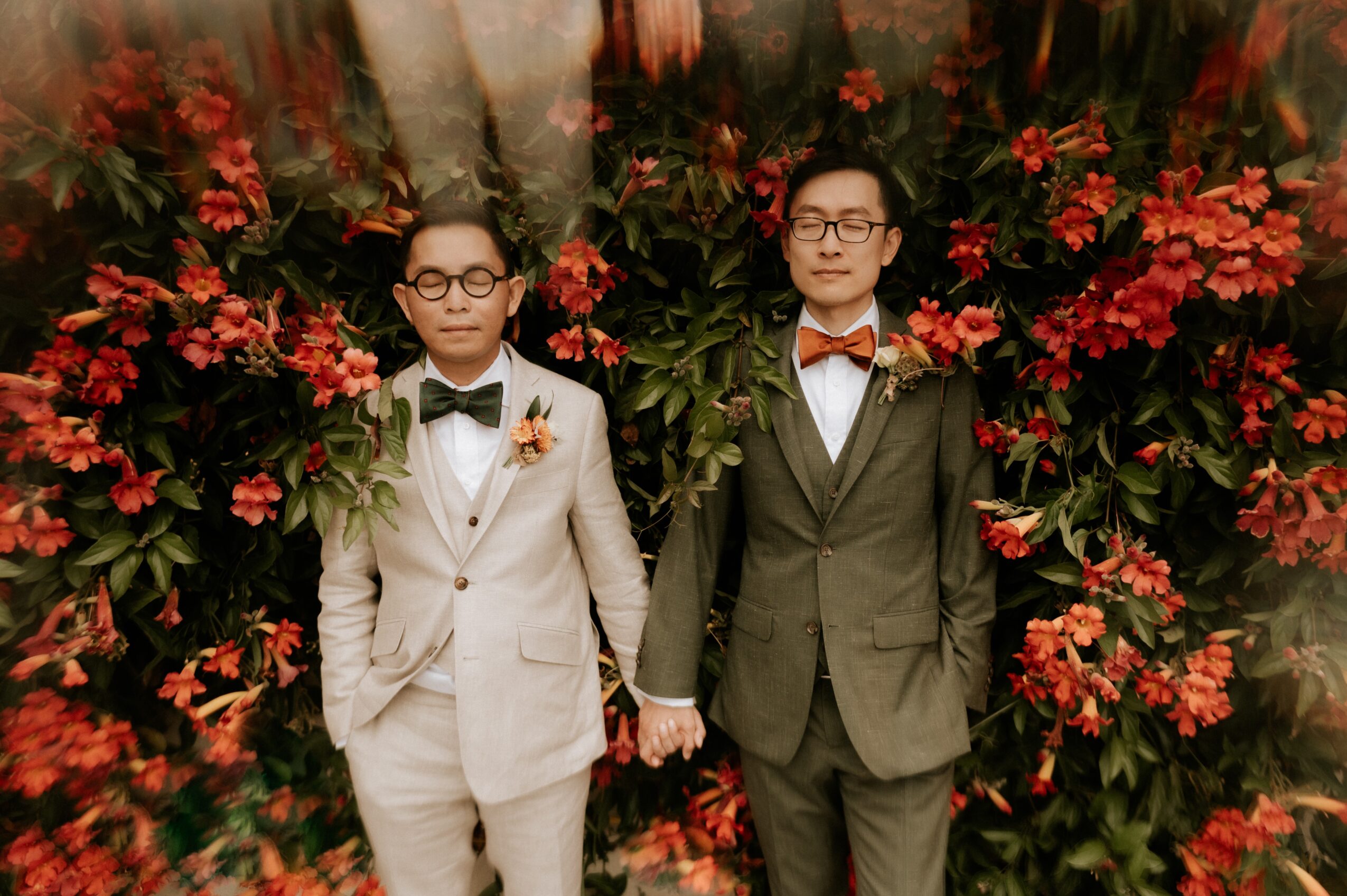 Two men in suits holding hands, standing in front of a floral backdrop with red flowers, both wearing glasses and bow ties.
