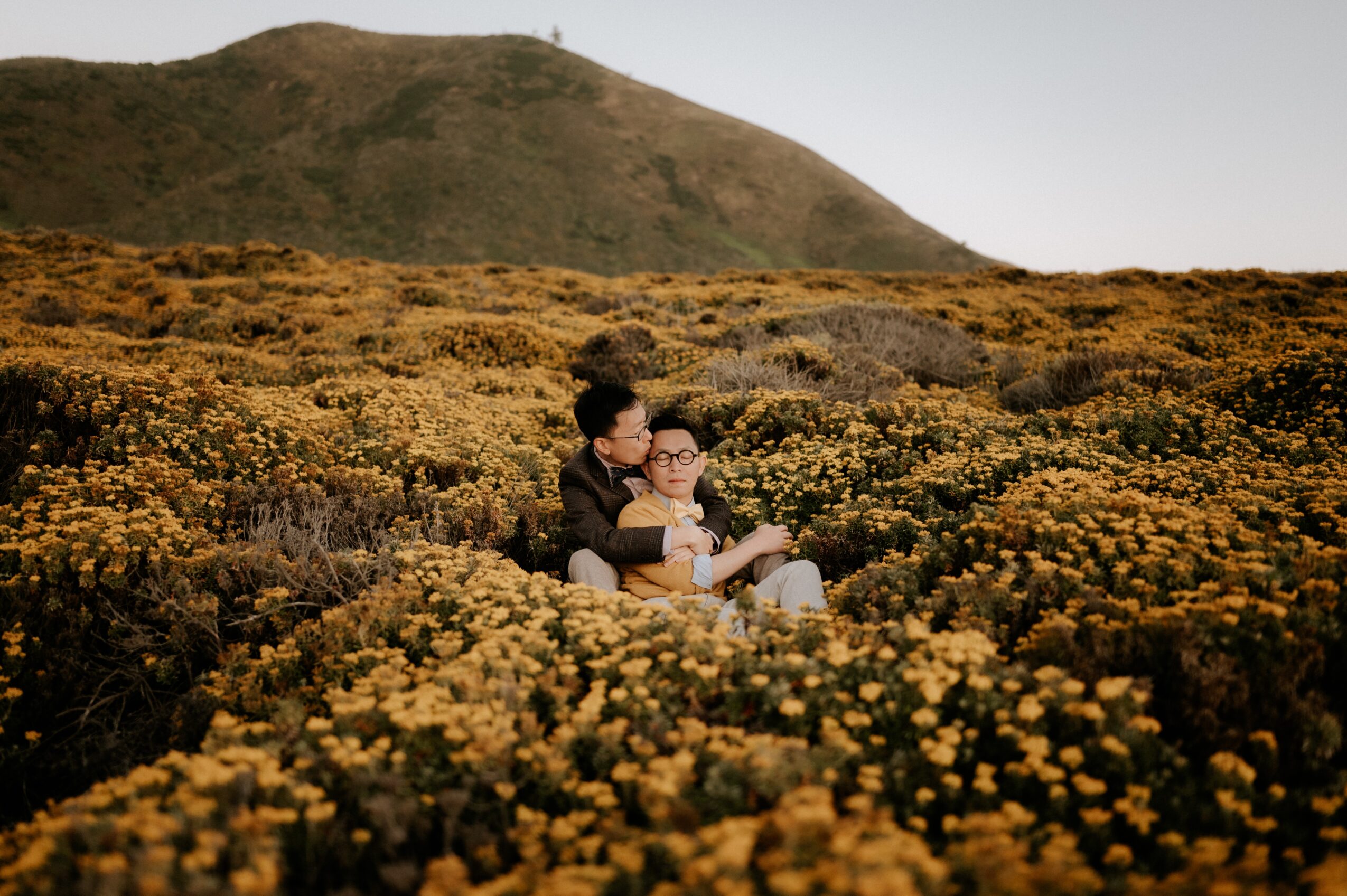 Two people sitting in a field of yellow flowers with a mountain in the background.