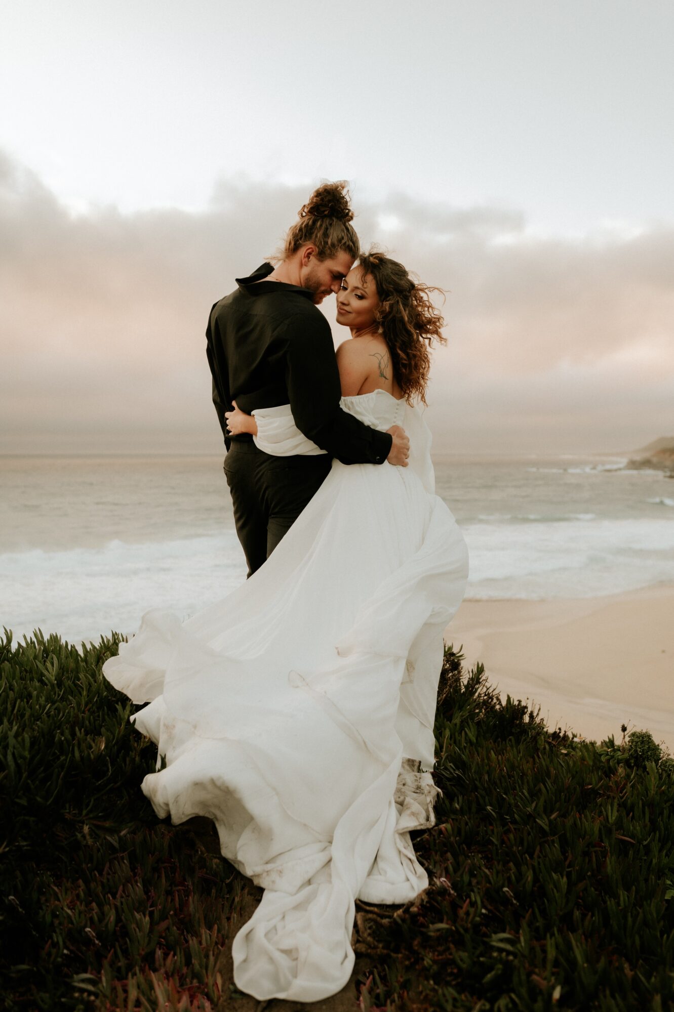 A couple in wedding attire standing on a beach, embracing each other with ocean and cloudy sky in background.