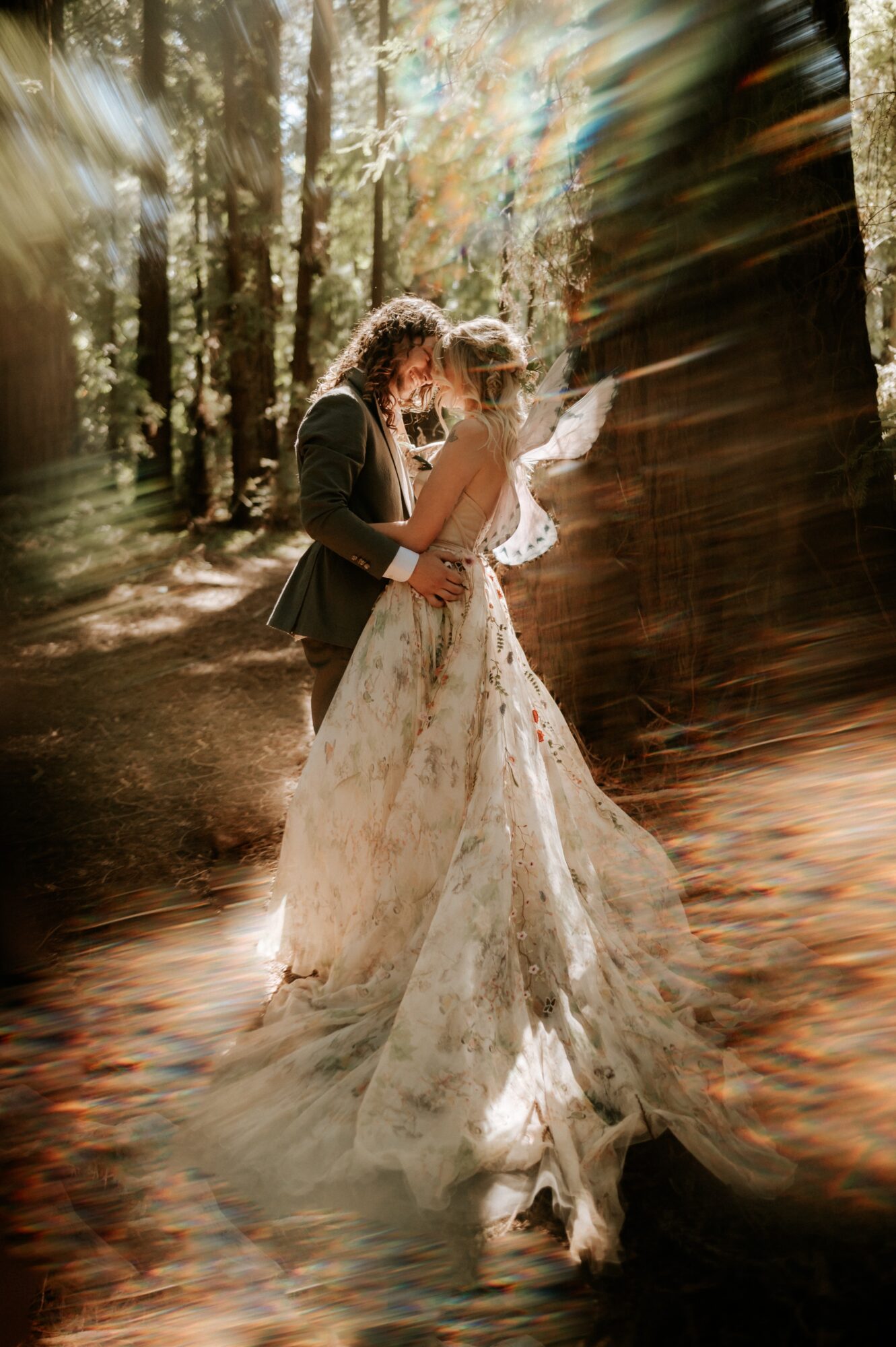 A couple in wedding attire embracing in a sunlit forest, with light rays and blurred background.