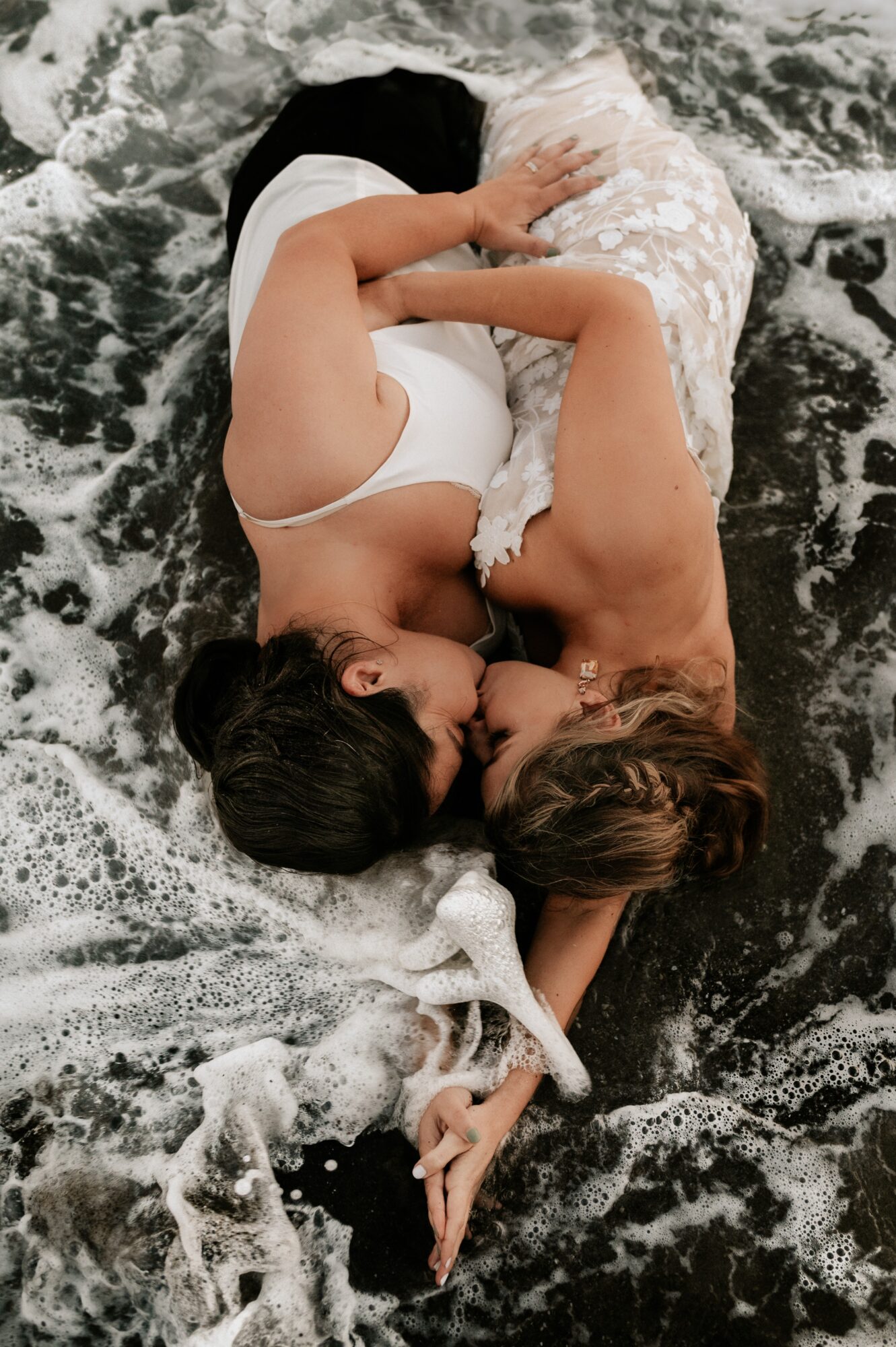 Two women lying on wet rocks near water, embracing and touching each other, with dark hair and light clothing.