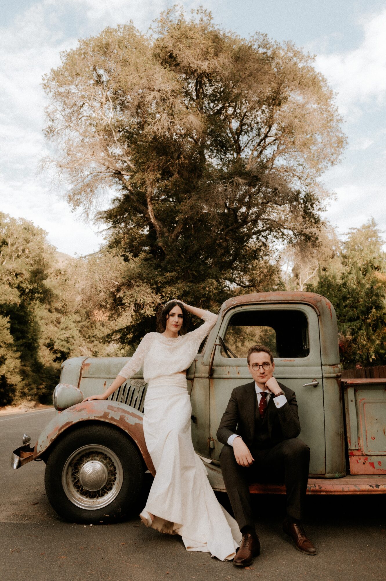 A woman in a white dress and a man in a suit pose beside a vintage truck outdoors with a large tree in the background.