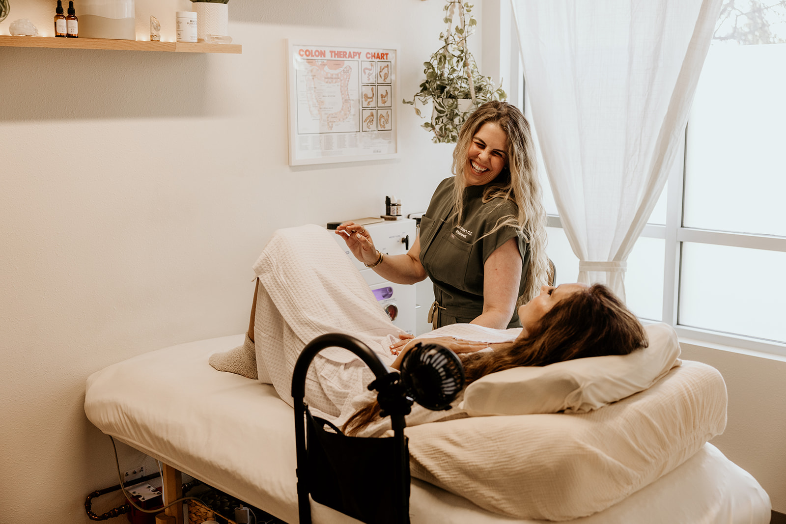 A woman smiling at a patient lying in a hospital bed near a window with white curtains.