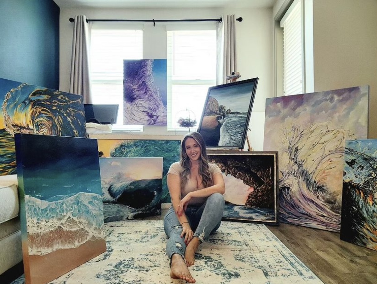Person sitting on a rug surrounded by multiple landscape paintings and framed artwork in a bright room.