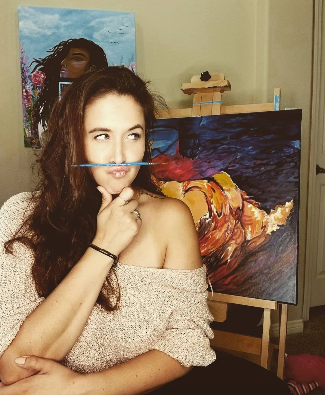 Young woman with long dark hair, wearing an off-shoulder sweater, standing in front of paintings, touching her chin thoughtfully.