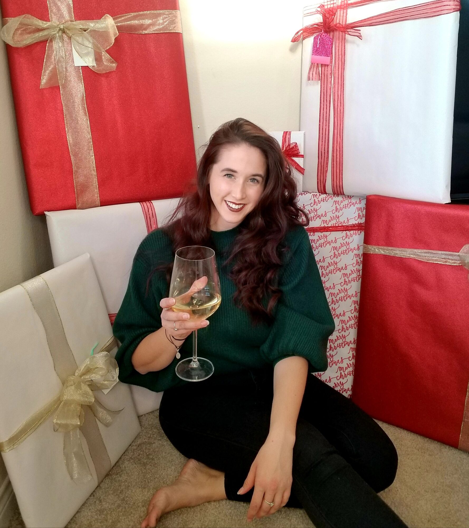 Woman sitting on floor holding a glass of wine, surrounded by large wrapped presents, smiling.