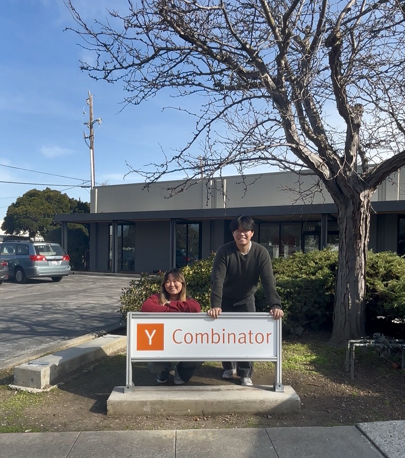 A man and a girl stand behind a sign that reads 'Y Combinaor' outdoors, with a tree and building in the background.