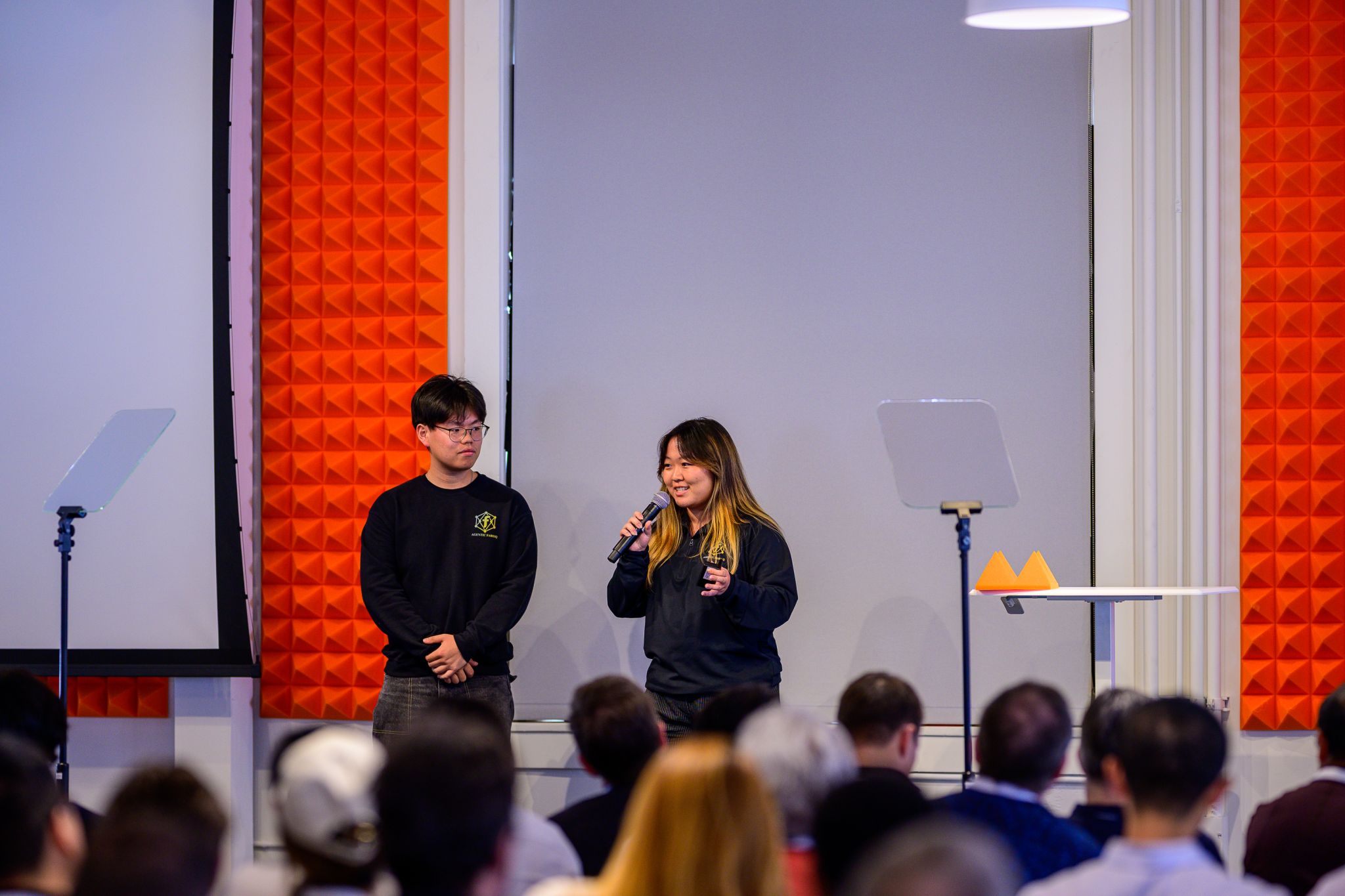 Two people stand on stage speaking to an audience, with orange soundproof panels behind them, and a white wall in the background.