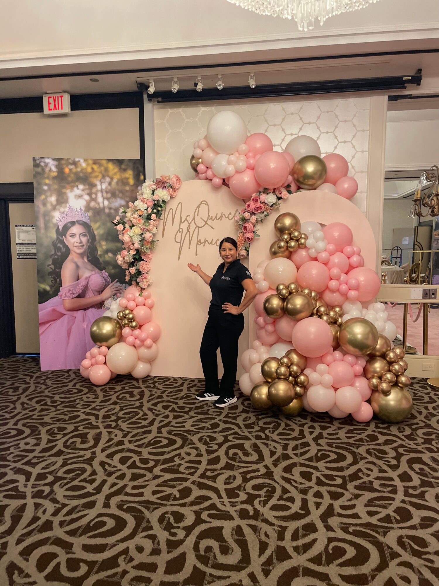 Person standing in front of pink, white, and gold balloon arch and floral display at event, with a backdrop and poster nearby.